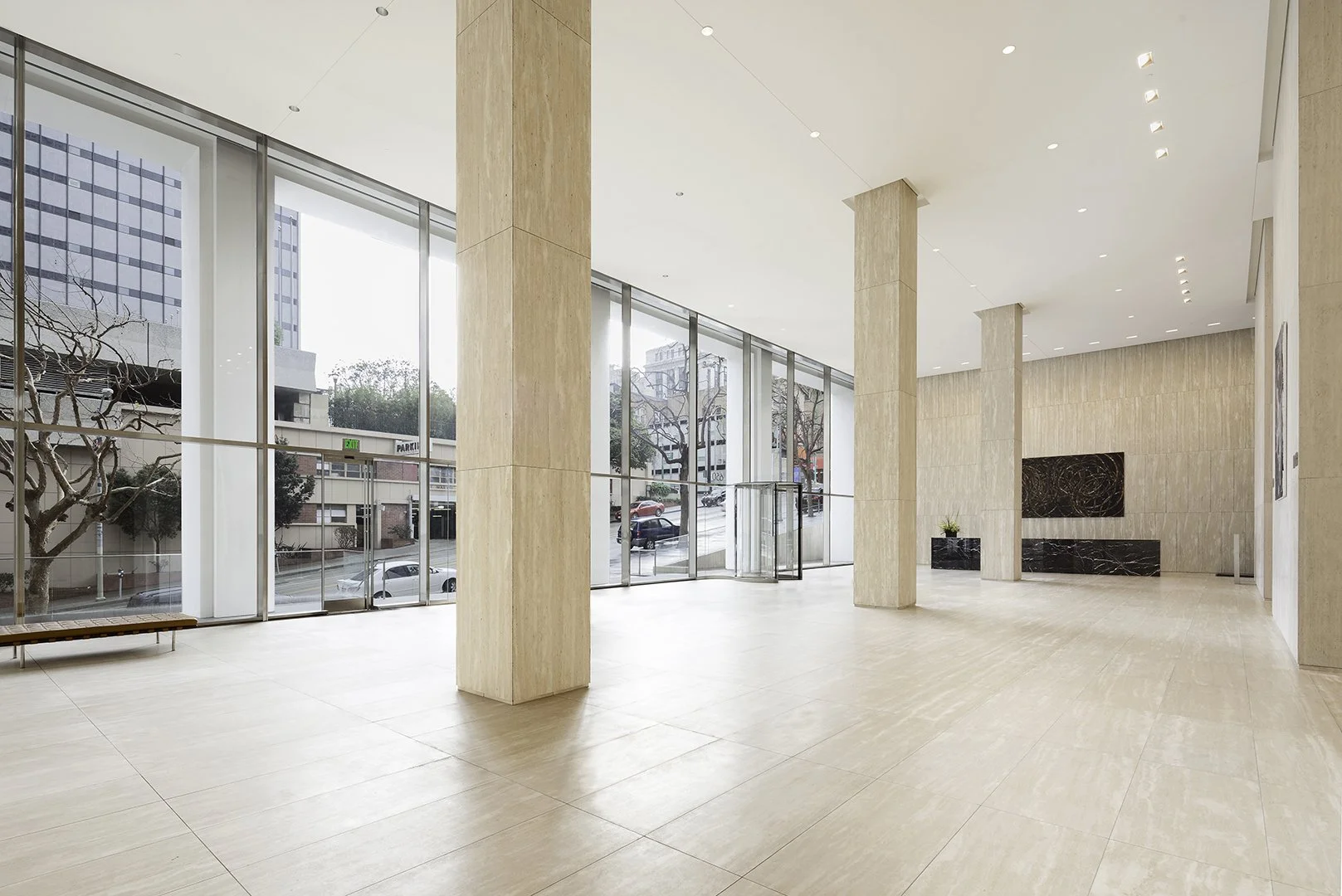 Empty lobby or reception area with large windows, beige tiled floors, and beige marble columns. Light-colored walls with modern artwork, a bench near the window, and a potted plant on a black marble stand. Outside, parked cars and trees are visible t