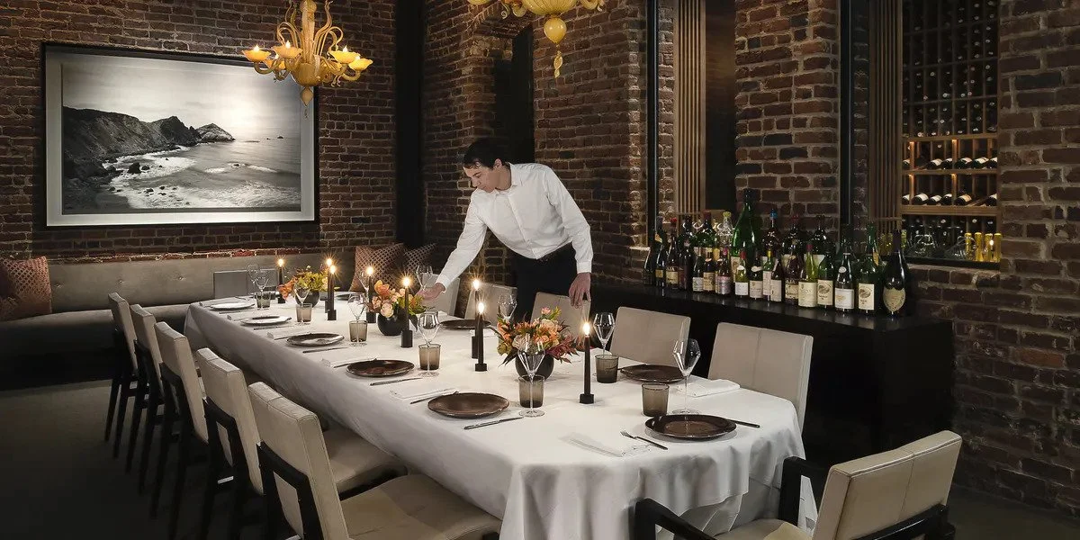 A person setting a long dining table with white tablecloth in a restaurant with brick walls, artwork, and wine bottles, decorated with candles and flower arrangements.