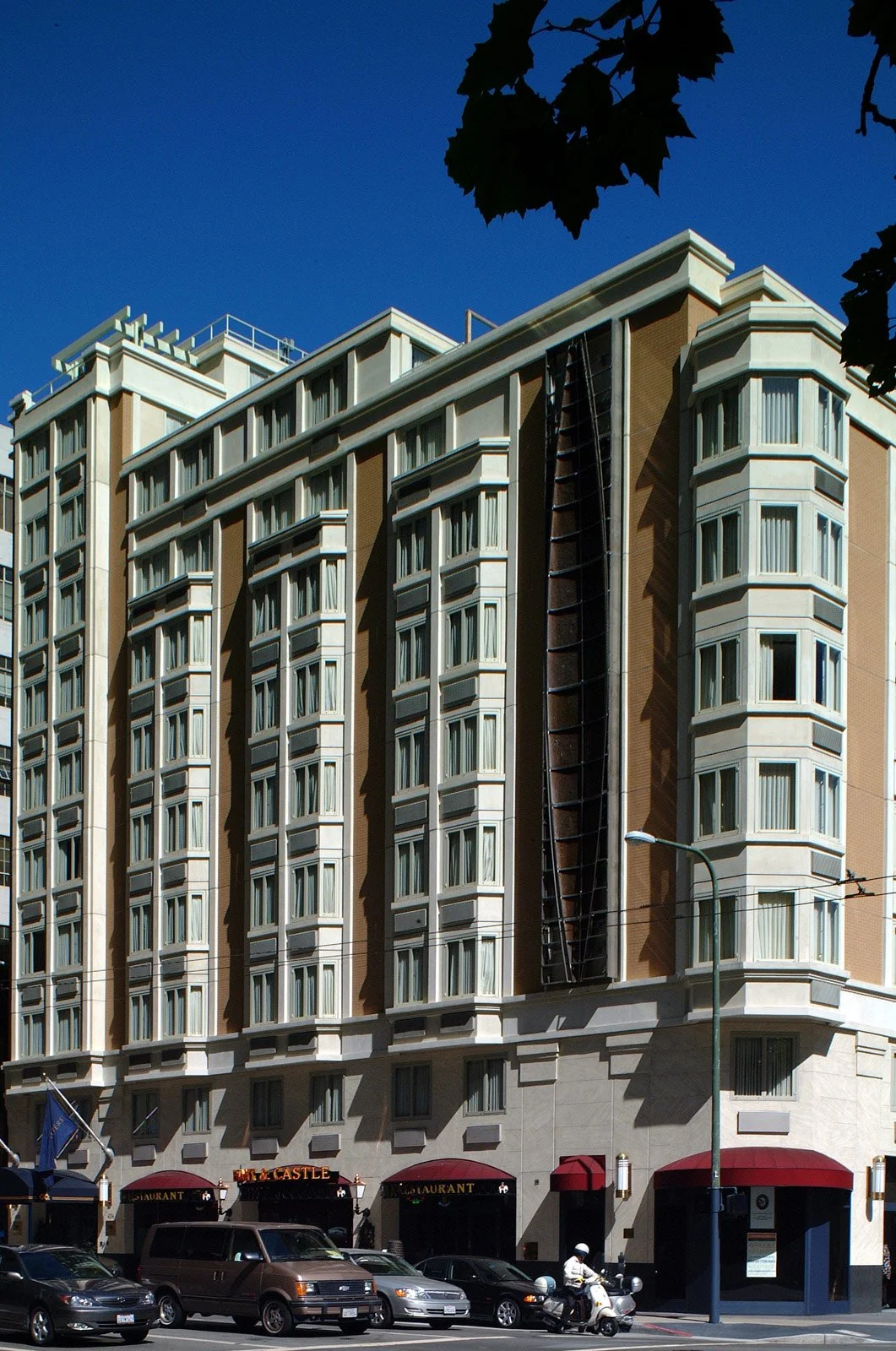 A tall multi-story building with beige and white exterior, many windows, a fire escape ladder on the front, and red awnings at the ground level, with parked cars and a motorcycle in front.