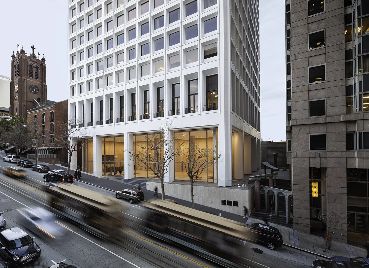 Modern white office building on city street with moving light trail from passing vehicle, trees, and neighboring buildings.