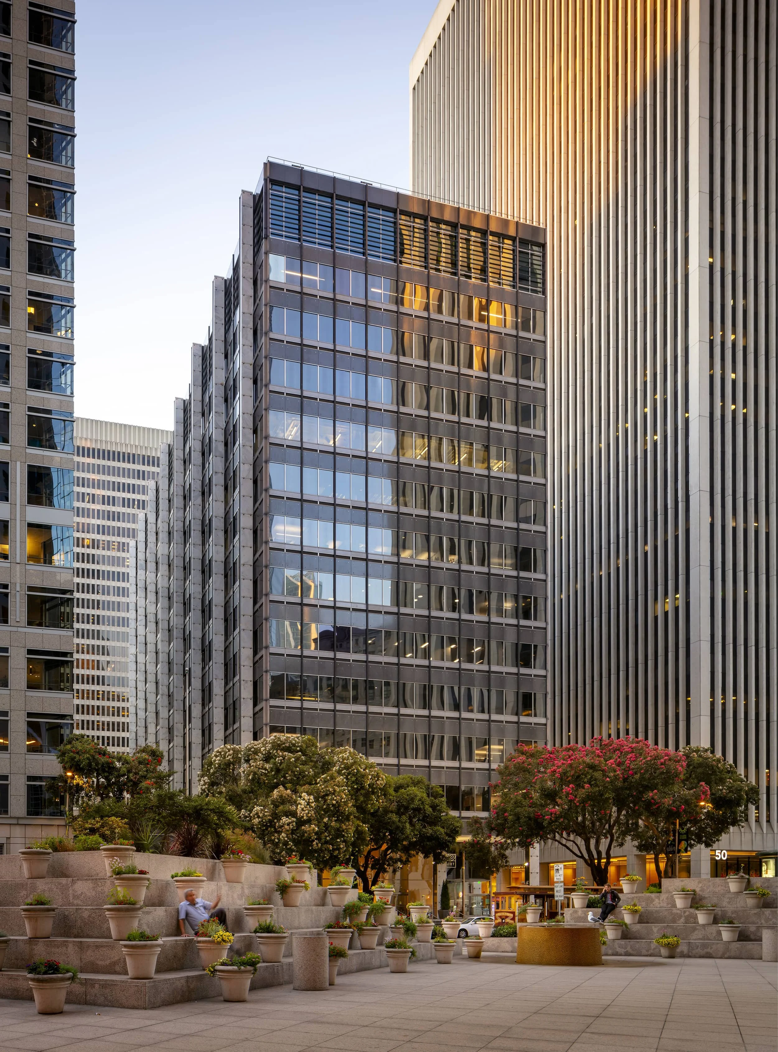 Cityscape with tall glass and steel office buildings, landscaped plaza with trees and potted plants, and a person sitting on steps in the foreground at sunset.