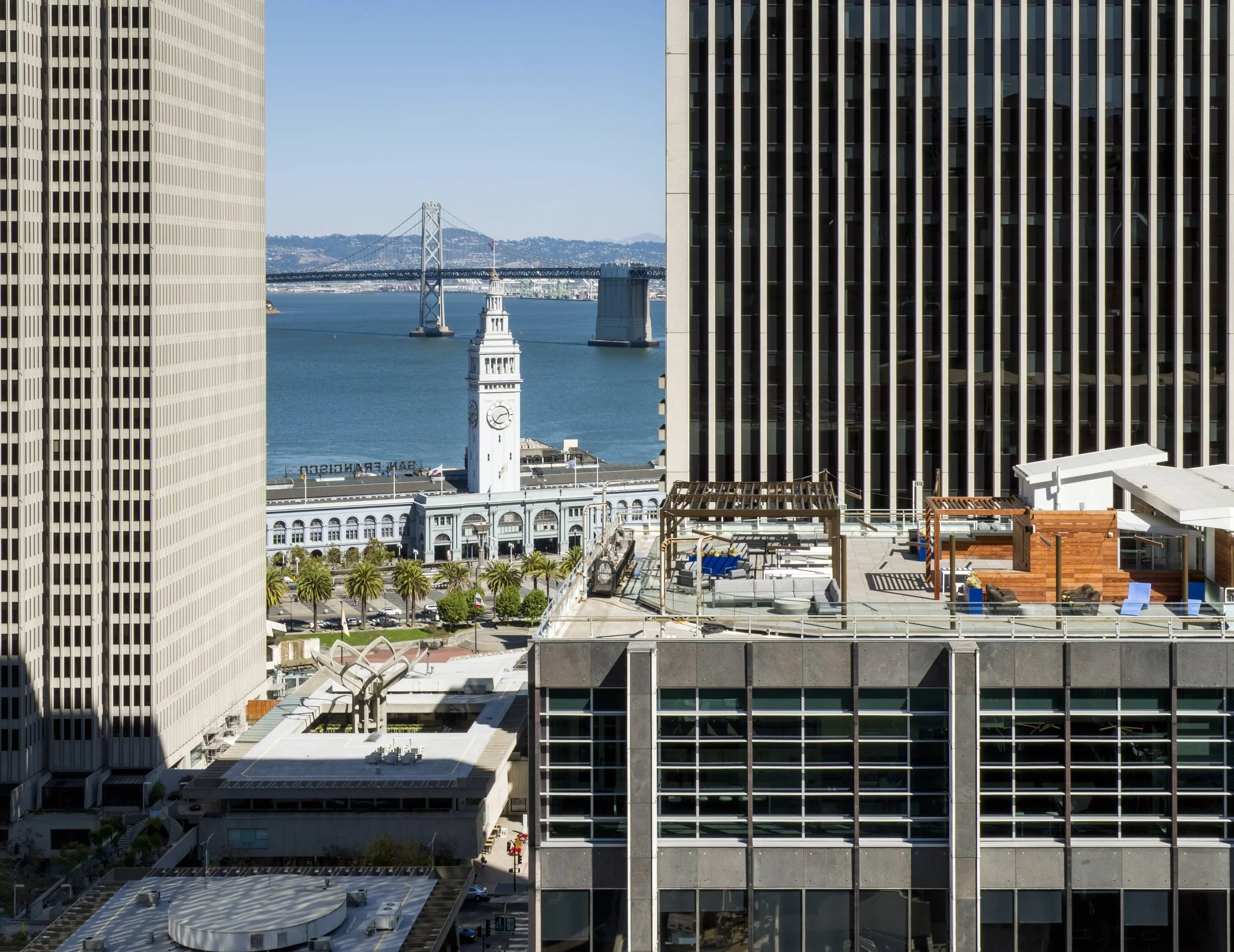 View of San Francisco Bay framed by tall office buildings, with the Ferry Building and the Bay Bridge visible in the distance on a clear day.