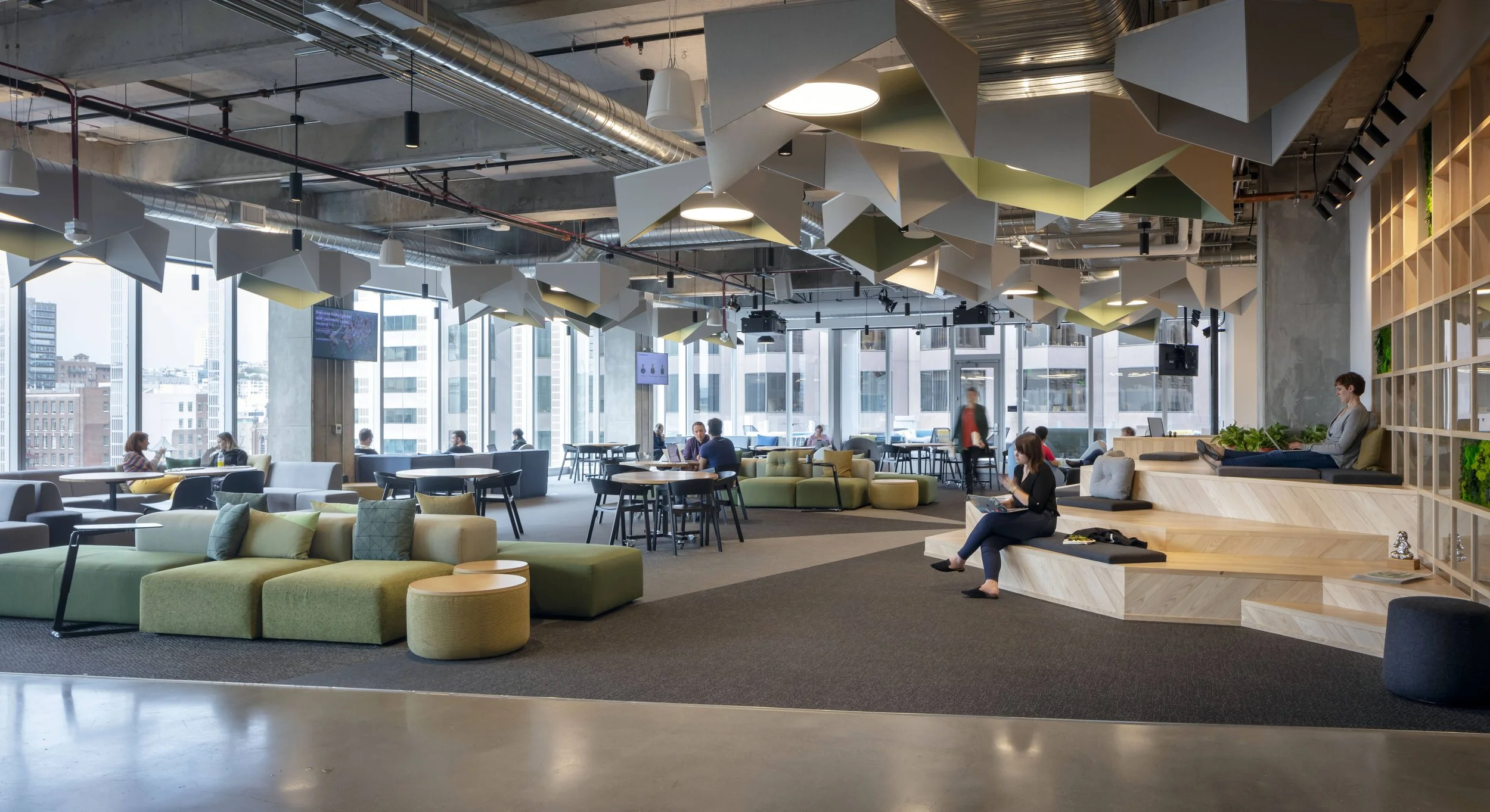 A modern, open-concept coworking space with large windows providing city views. The room features green and gray lounge seating, black tables, and a tiered wooden platform where a woman is sitting. There are geometric ceiling acoustic panels and plan