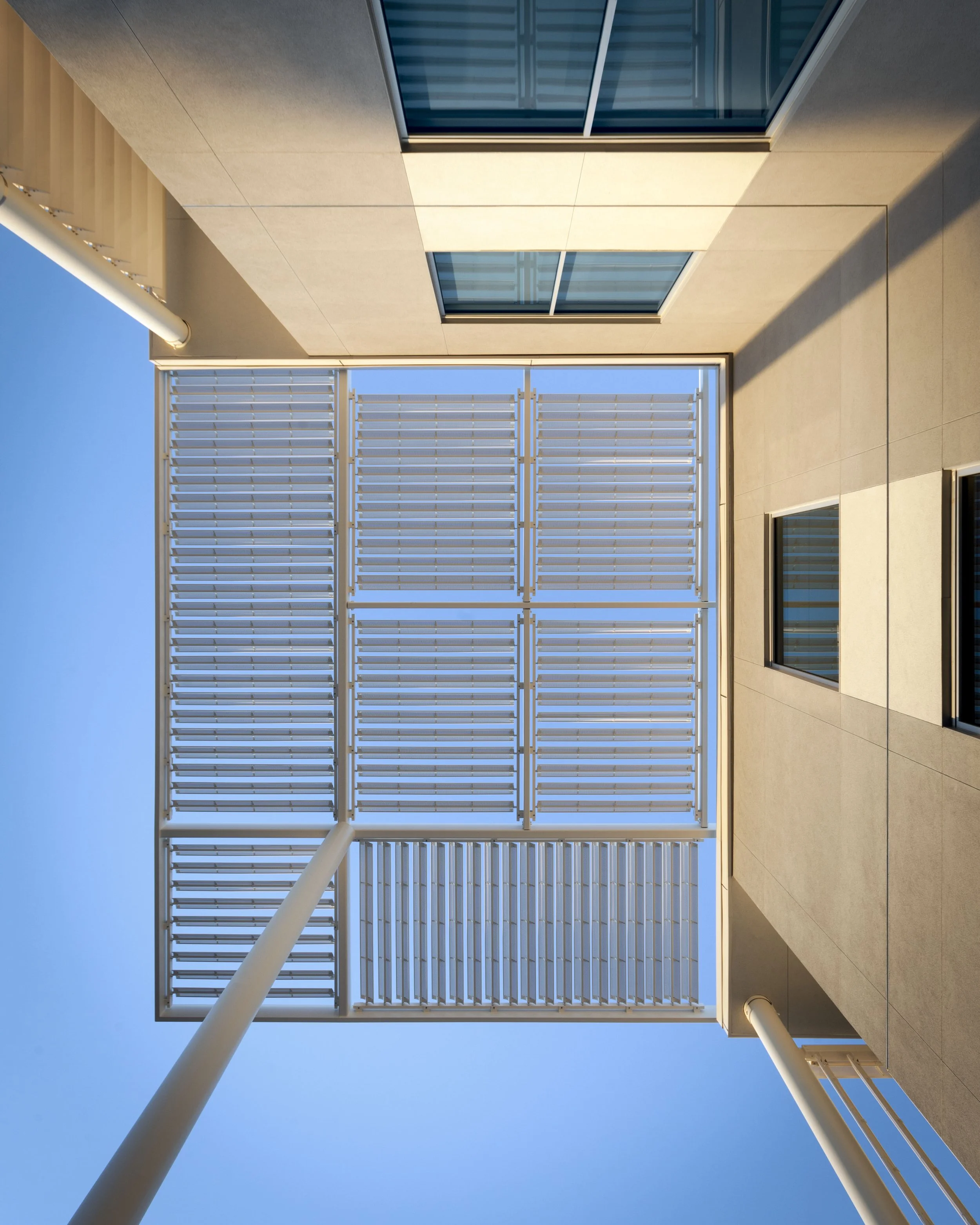 Looking up at the sky through a modern building's rooftop solar panel structure with metal slats, supported by poles, against a clear blue sky.
