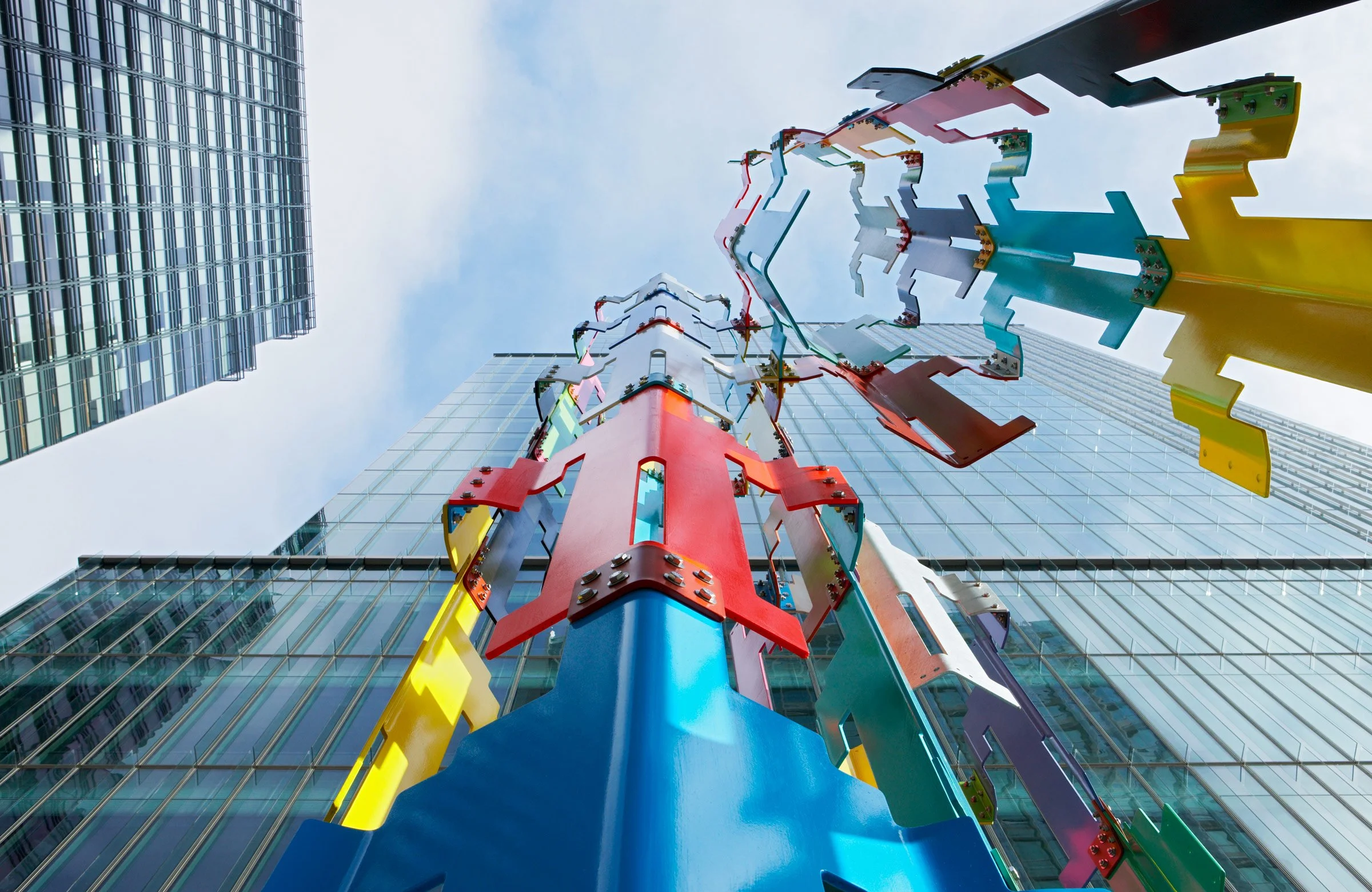 Colorful abstract sculpture mounted on a building facade, seen from below, with surrounding skyscrapers and a cloudy sky.