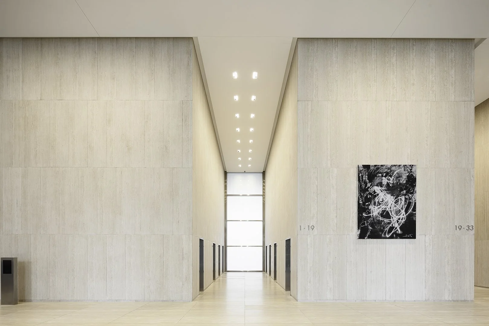 Modern art gallery with beige walls, a narrow corridor, and an abstract black and white painting on the right wall.