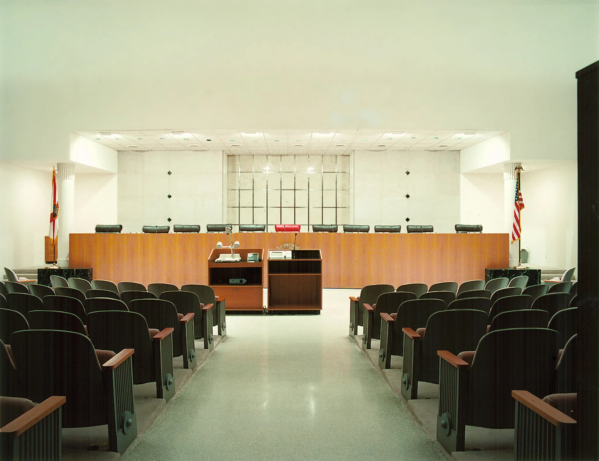 Empty courtroom with a wooden judge's bench, chairs for the judge and attorneys, American flags on either side, and rows of audience seats.
