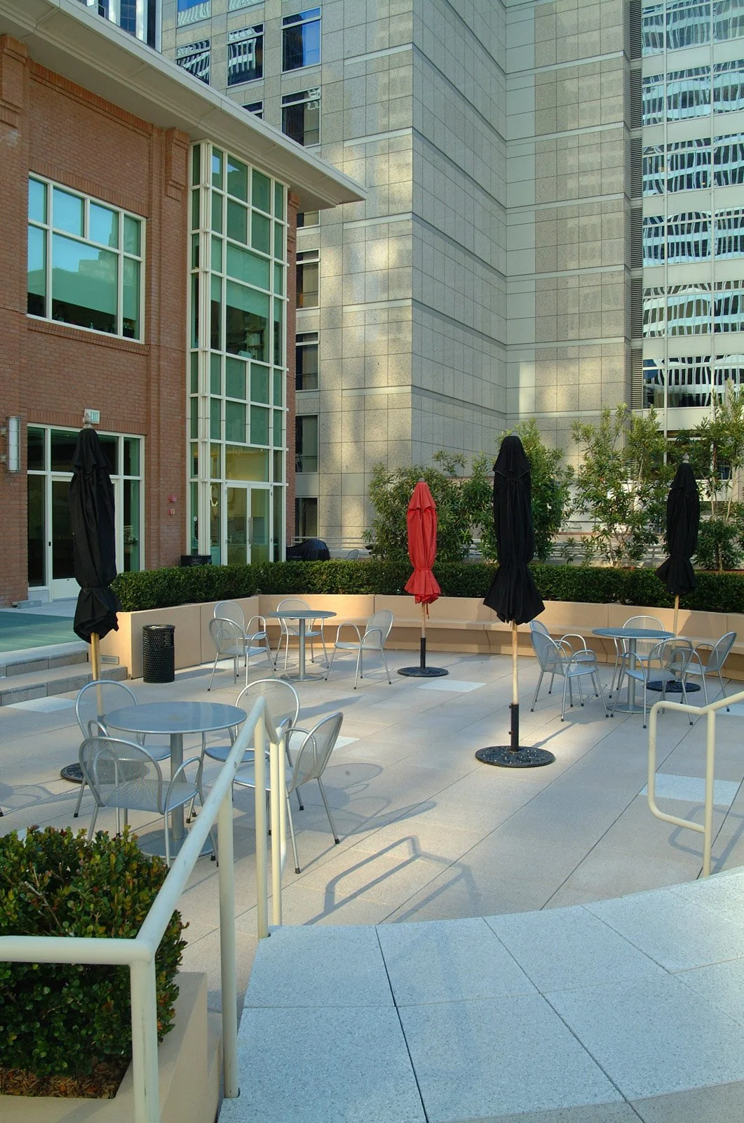 An outdoor patio area with round tables and metal chairs, some with closed black and red umbrellas, surrounded by modern high-rise buildings in an urban setting.