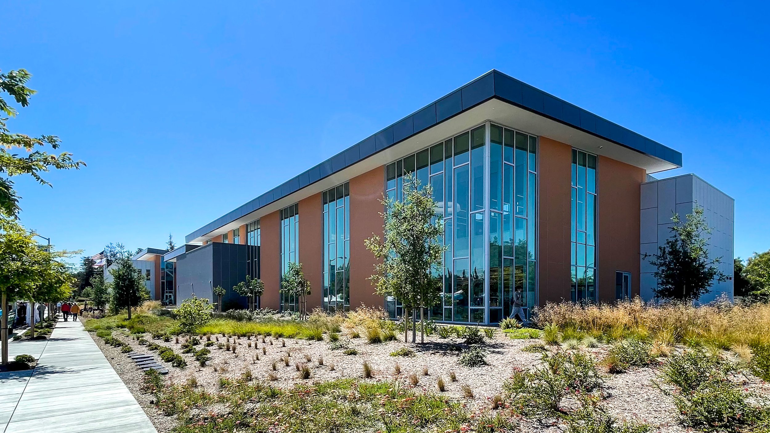 Modern building with large glass windows, surrounded by landscaping and trees, under a clear blue sky.