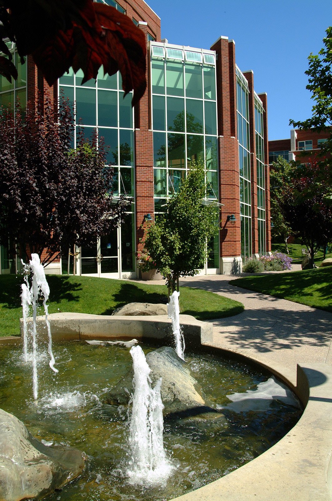A courtyard with a fountain, trees, and sidewalks in front of a modern building with large glass windows and brick walls.
