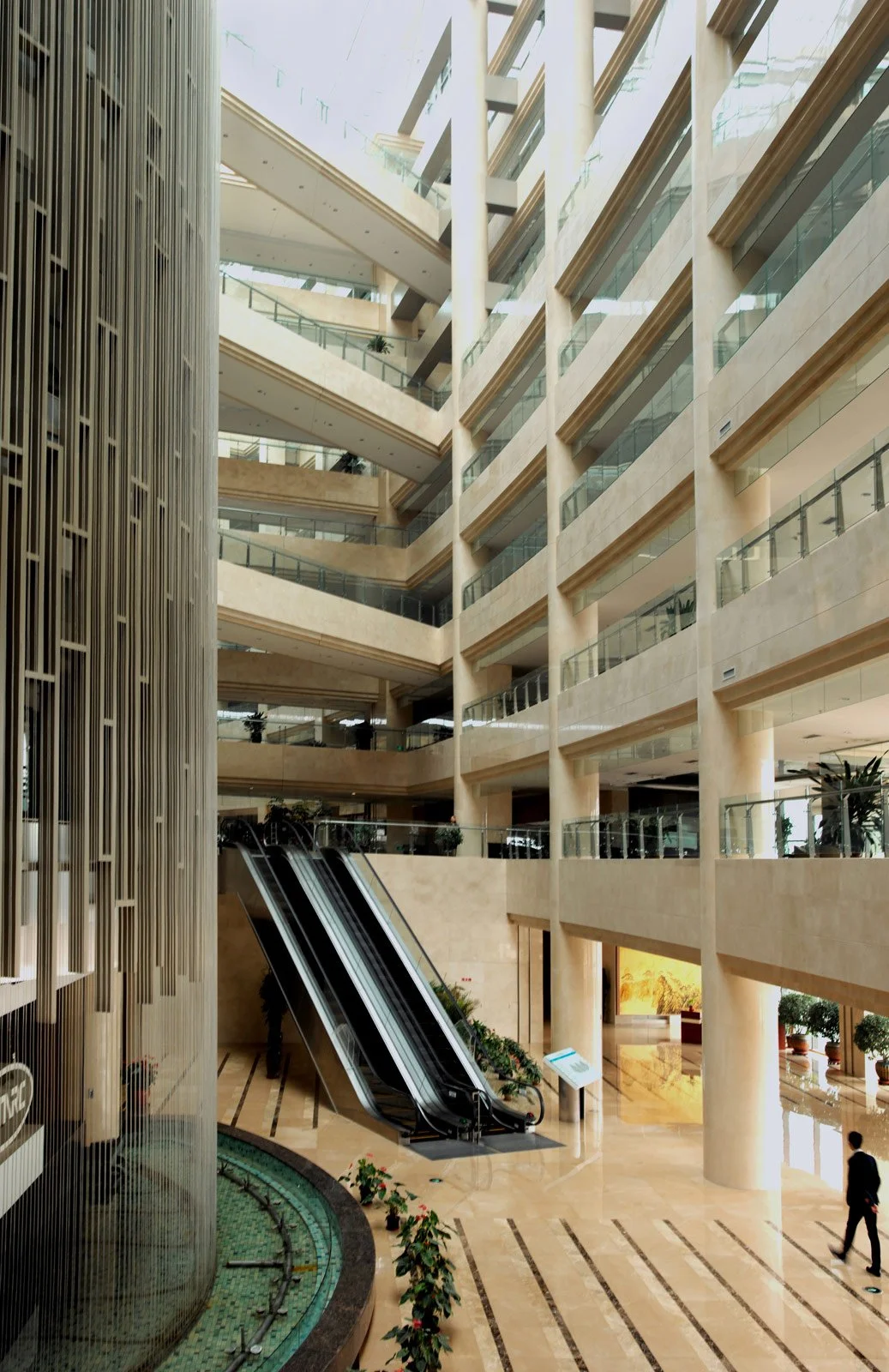 Interior view of a modern multi-story lobby with beige walls, glass railings, escalators, and a decorative fountain with plants.