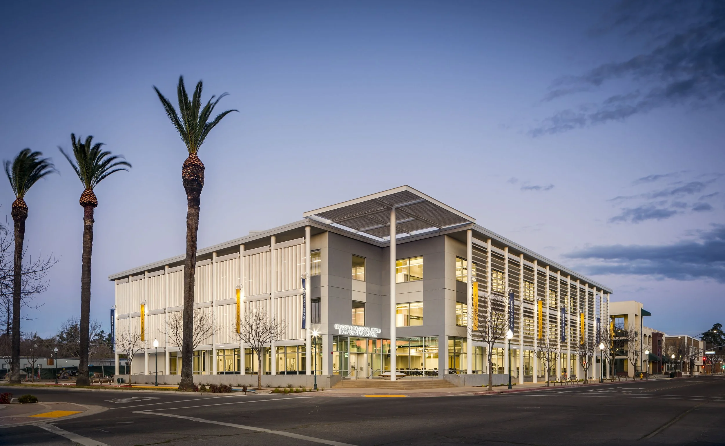Modern multi-story building with large windows, surrounded by palm trees and empty street, during dusk.