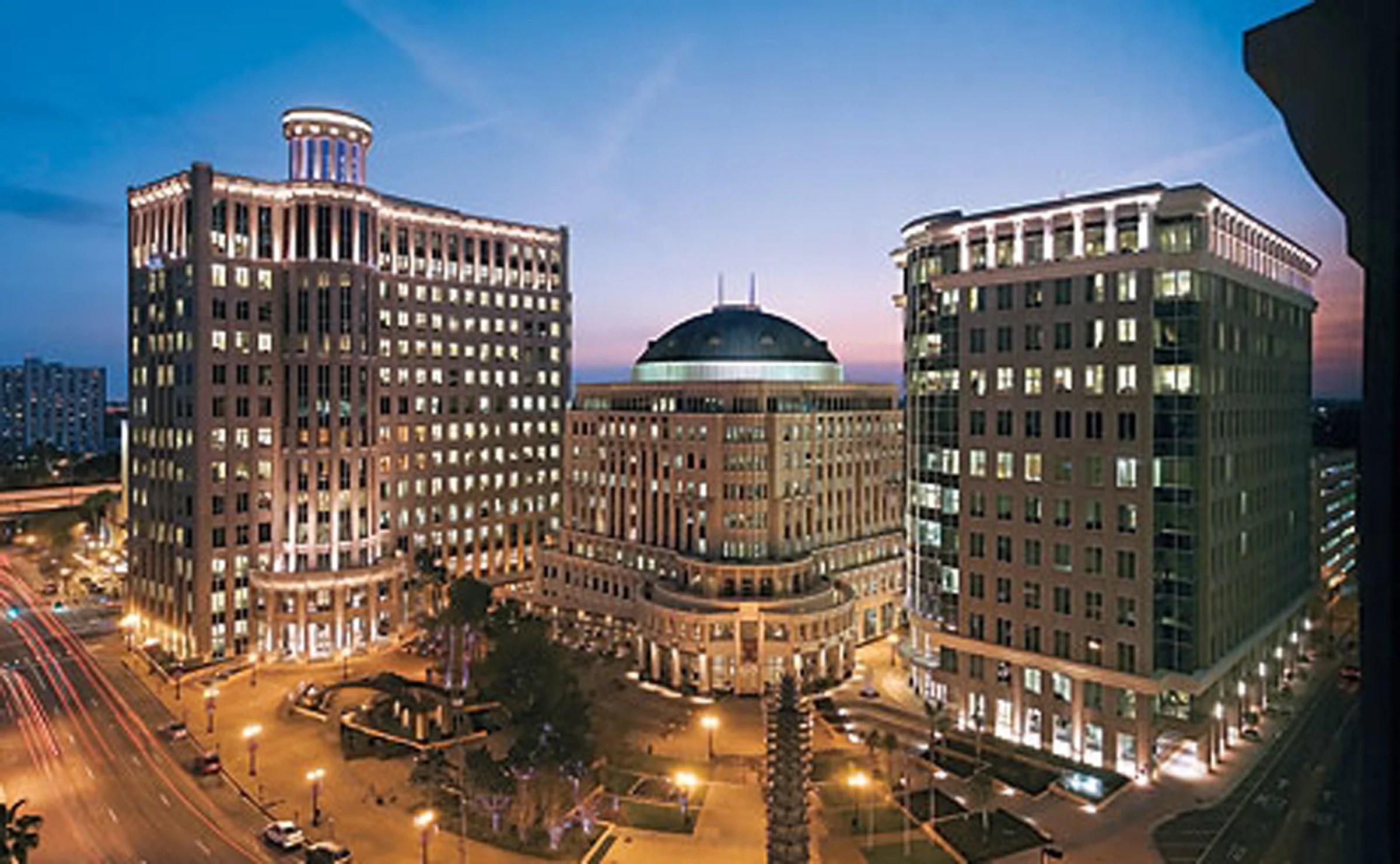 Nighttime cityscape featuring three tall buildings with illuminated windows, a central building with a rounded roof, and a busy street with streaks of car lights below.