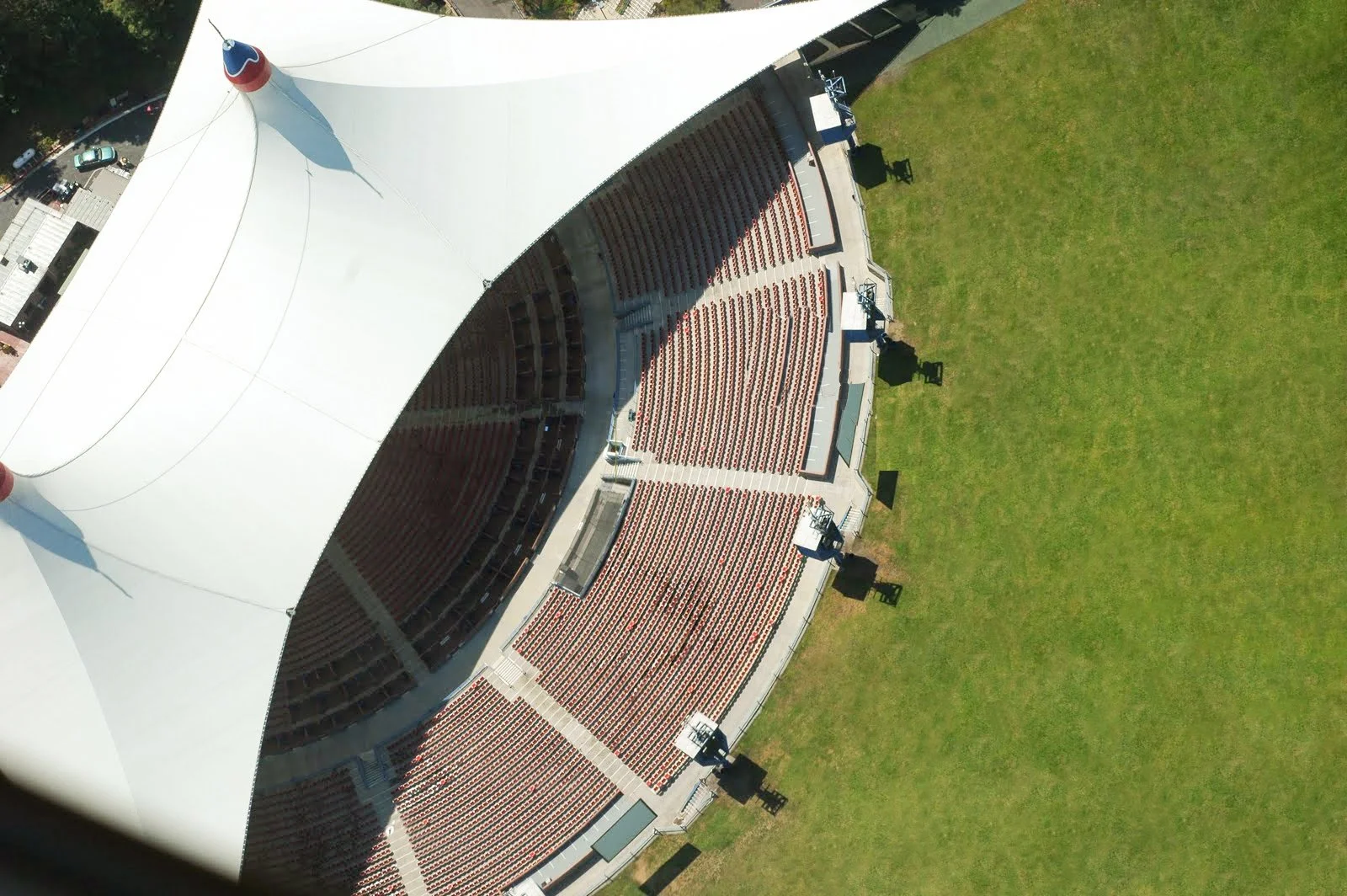 An aerial view of an outdoor amphitheater with red seats and a white tent roof, surrounded by a grassy area.