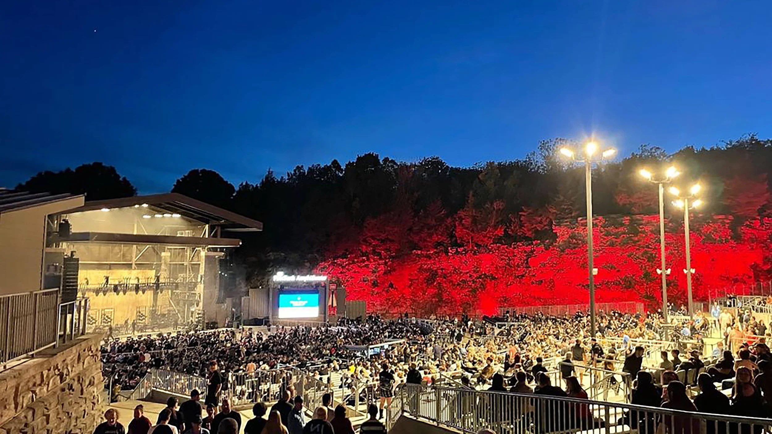 An outdoor amphitheater crowd at night with a stage on the left and a hillside with red trees in the background, illuminated by stage lights and streetlamps.