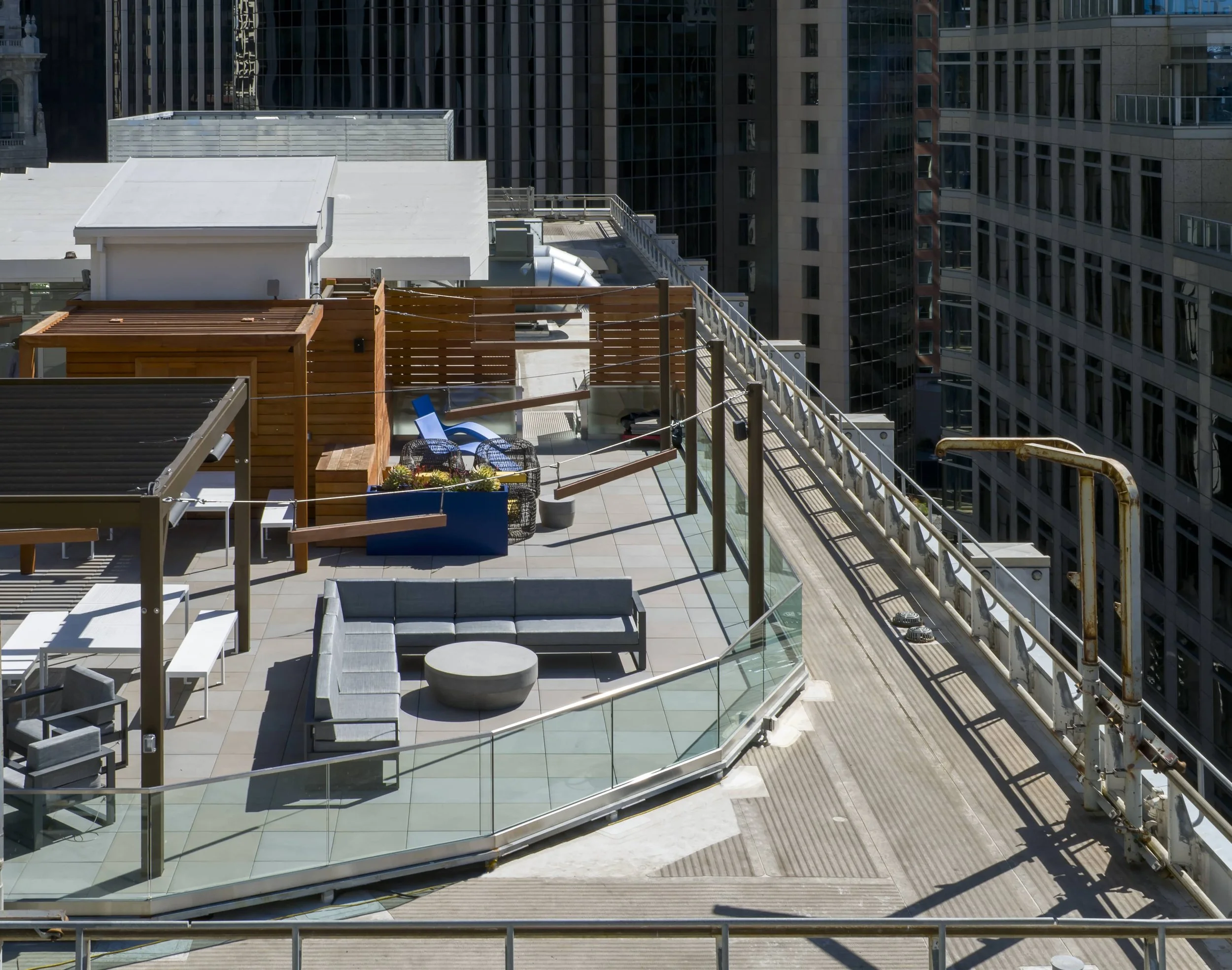 Rooftop terrace in an urban city with lounge furniture, patio chairs, a coffee table, and decorative plants, surrounded by glass railings and high-rise buildings.