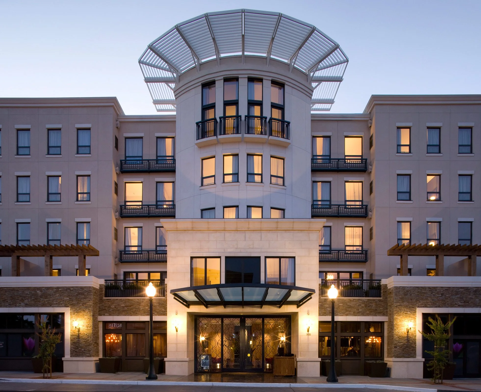 Front view of a modern, multi-story hotel building during dusk, with lit windows, outdoor lamps, and a glass entrance door.