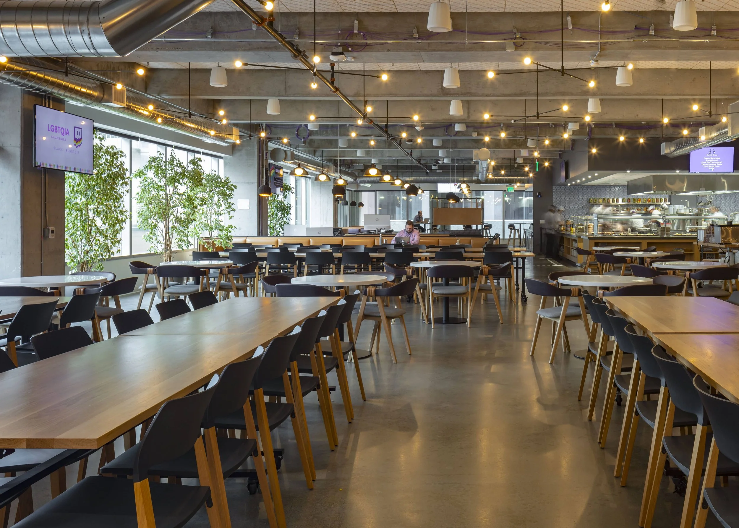 Interior of a modern, spacious restaurant with wooden tables and black chairs, large windows with green plants, and warm lighting fixtures hanging from the ceiling. A person is working at a table, and the open kitchen is visible in the background.
