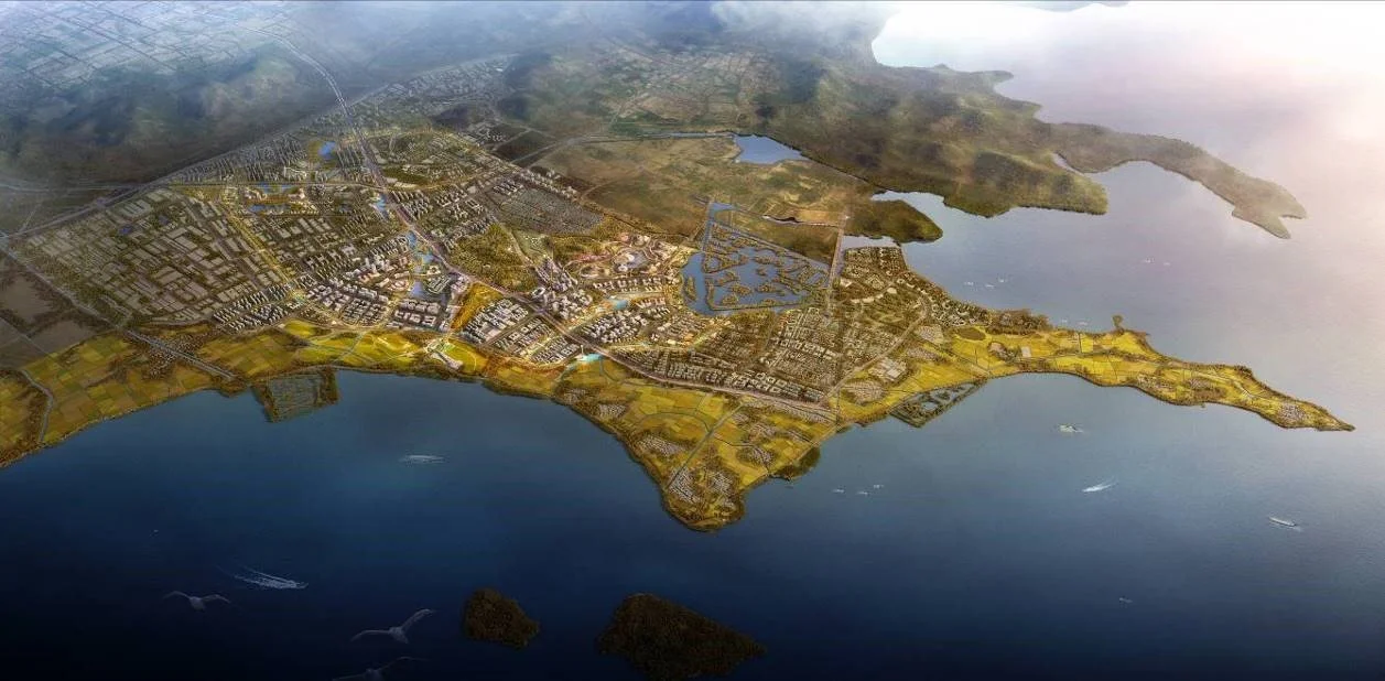 Aerial view of a coastal city with a mix of urban development, roads, water bodies, and green areas, along a shoreline with some boats on the water.