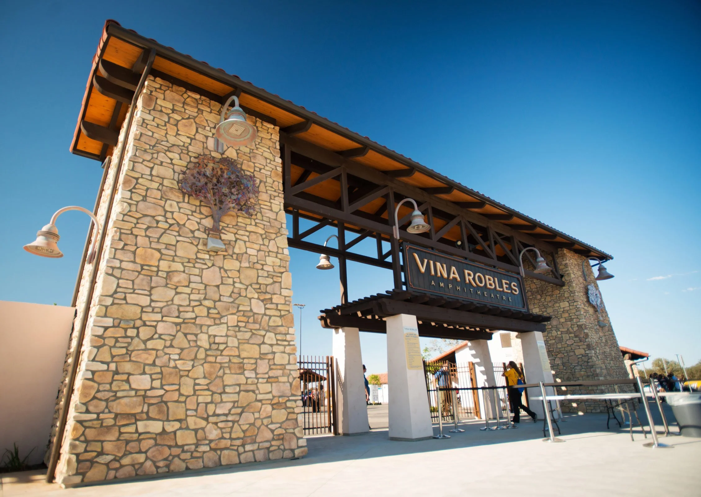 Exterior of Vina Robles Amphitheatre with stone and wood architecture, signage, and people entering.