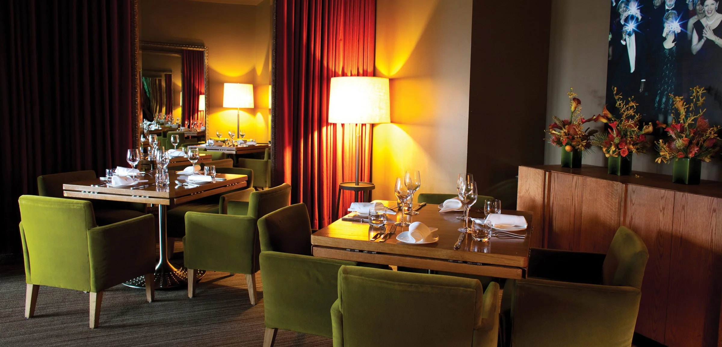 Elegant restaurant dining area with tables set with white napkins, glasses, and silverware, green velvet chairs, warm lighting from lamps, red curtains, and floral decorations.