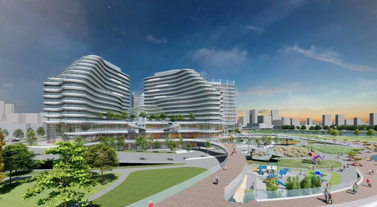 A modern urban development with two large, curvy glass buildings surrounded by landscaped park areas, walkways, and a children's playground under a partly cloudy sky at sunset.