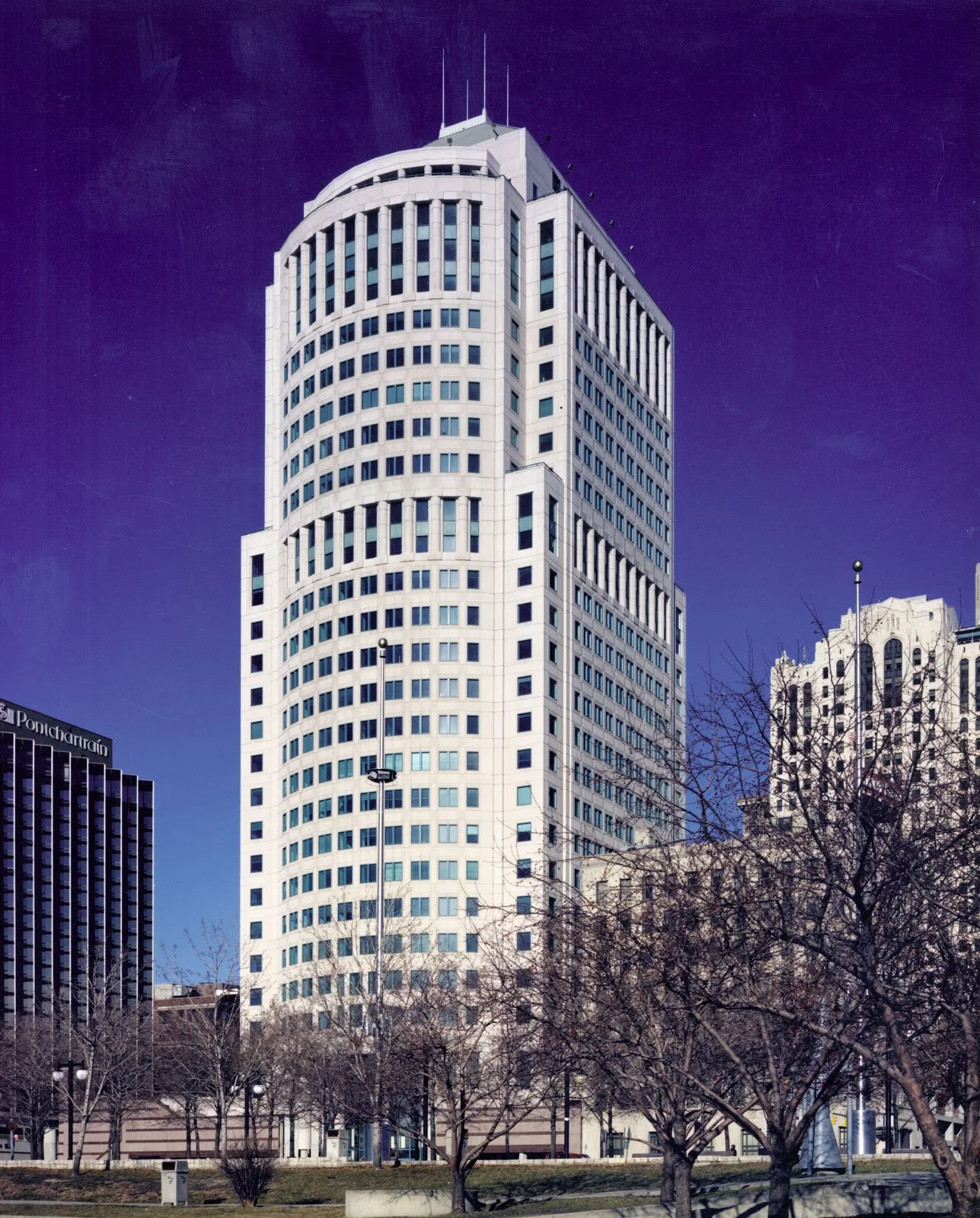 A tall white skyscraper with rounded edges and numerous windows, set against a dark purple sky, with leafless trees and surrounding buildings in an urban setting.