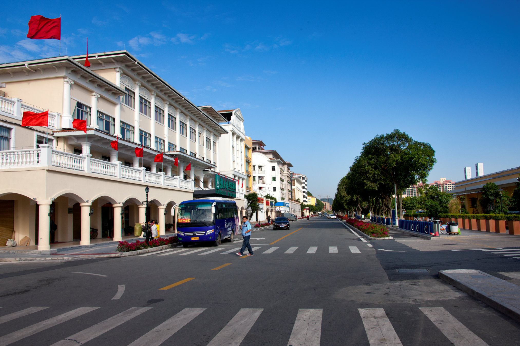 A city street with buildings on the left featuring red flags, a blue bus, and a person crossing the street; trees line the right side, and the sky is clear with minimal clouds.
