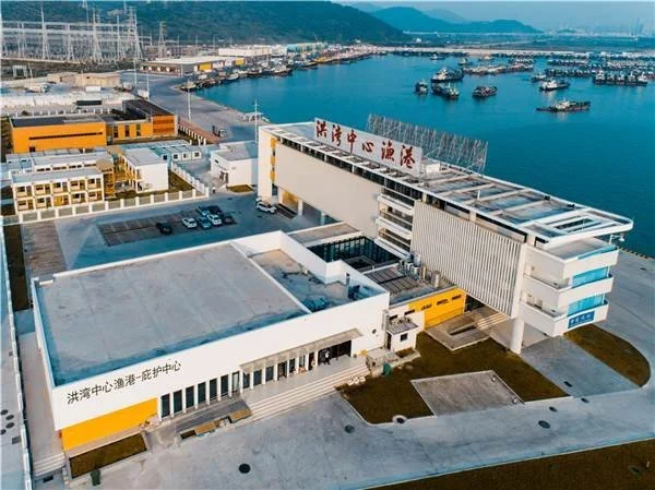 Aerial view of a modern port facility with buildings and a harbor filled with boats, mountains in the background.