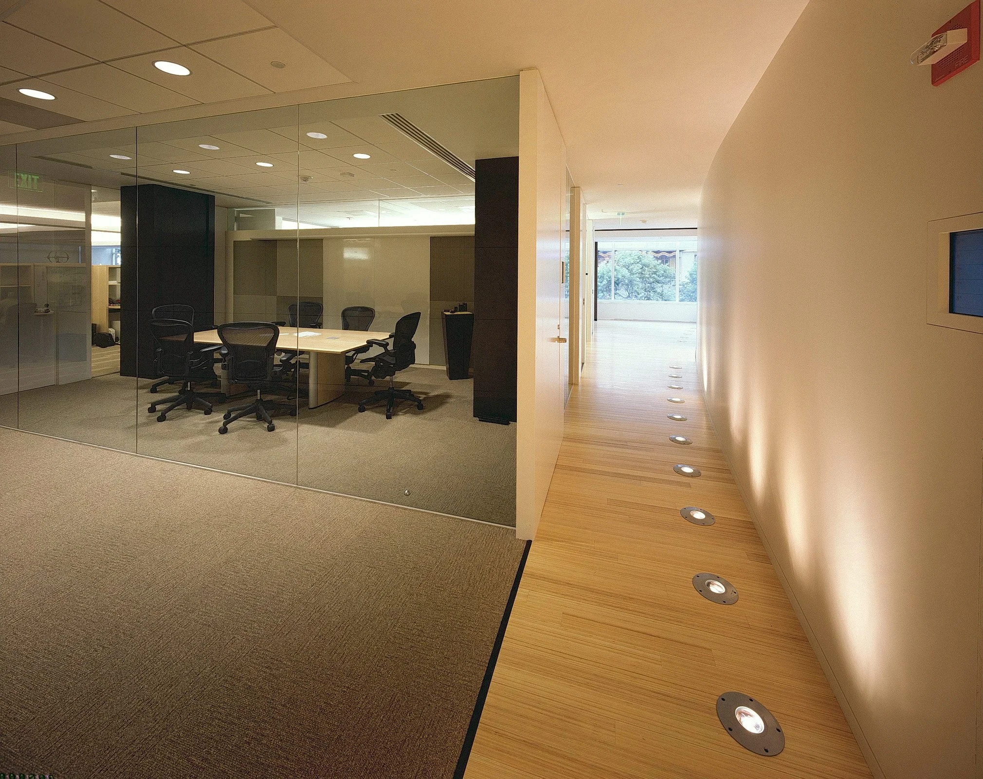 Empty conference room with a glass wall and six black office chairs around a central table, adjacent to a lit hallway with wooden flooring and small round ceiling lights.