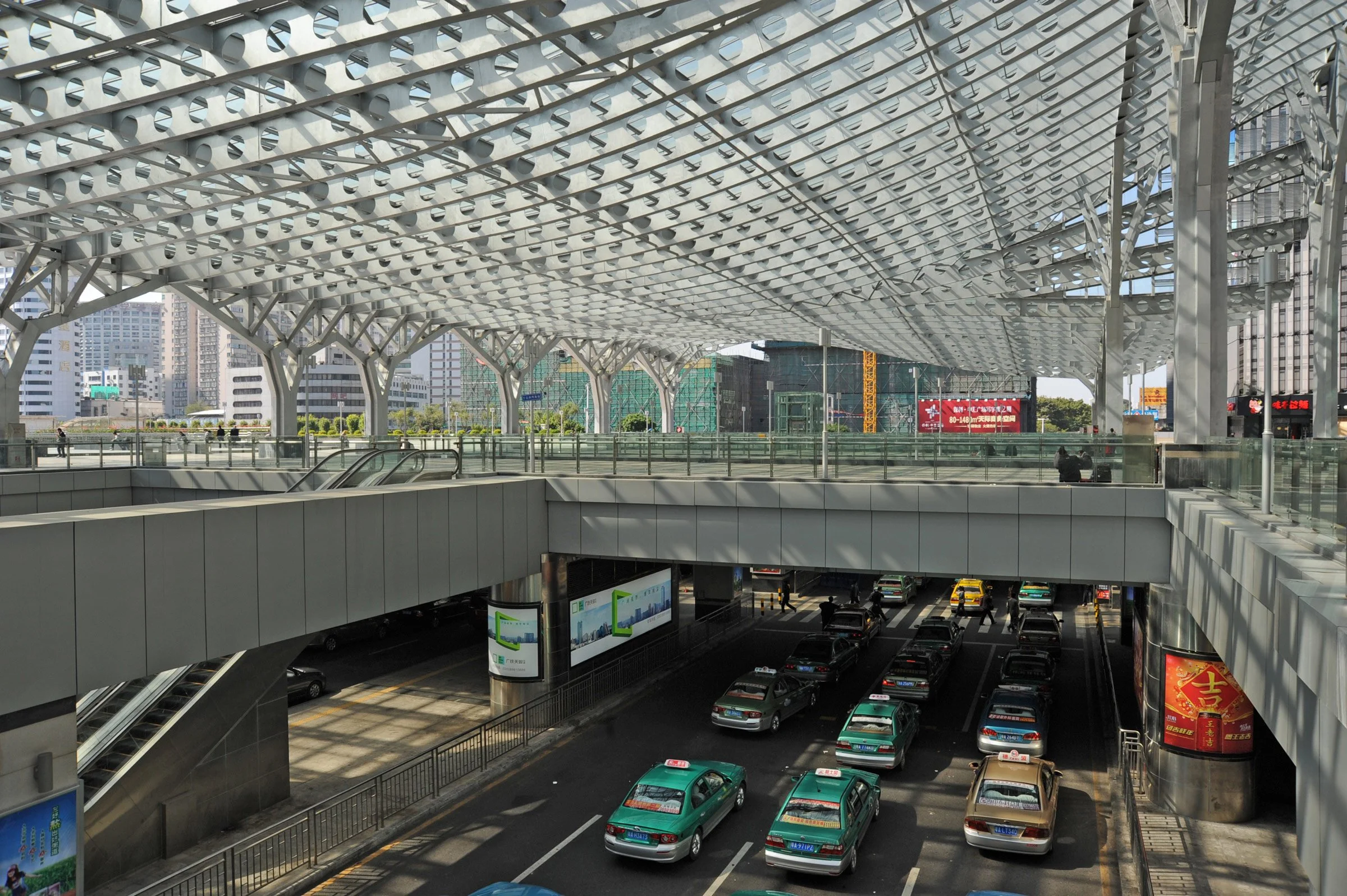 Modern urban transportation hub with a multi-level roof structure, multiple vehicles including taxis on a street, and buildings in the background.