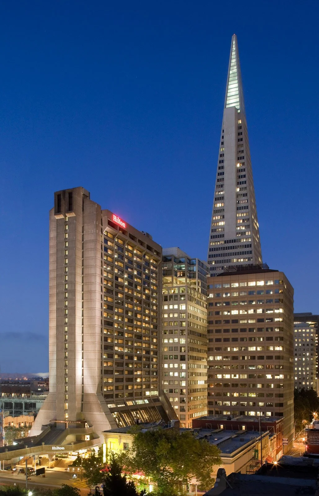 Nighttime cityscape featuring the Hilton hotel building with a unique architectural design and the Salesforce Tower with a sharp spire.