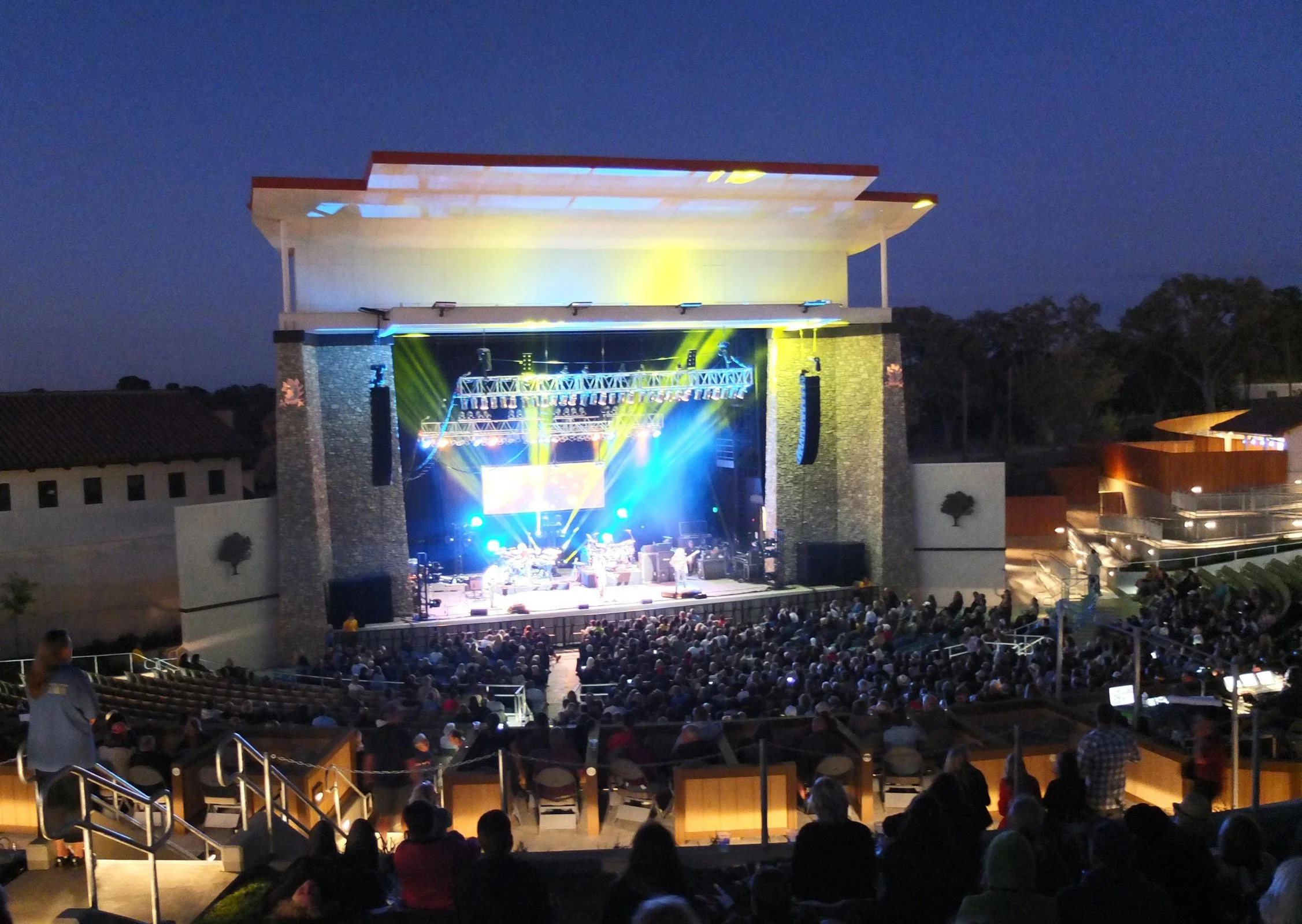 An outdoor concert at night with a large audience seated and a brightly lit stage featuring performers and colorful lights.