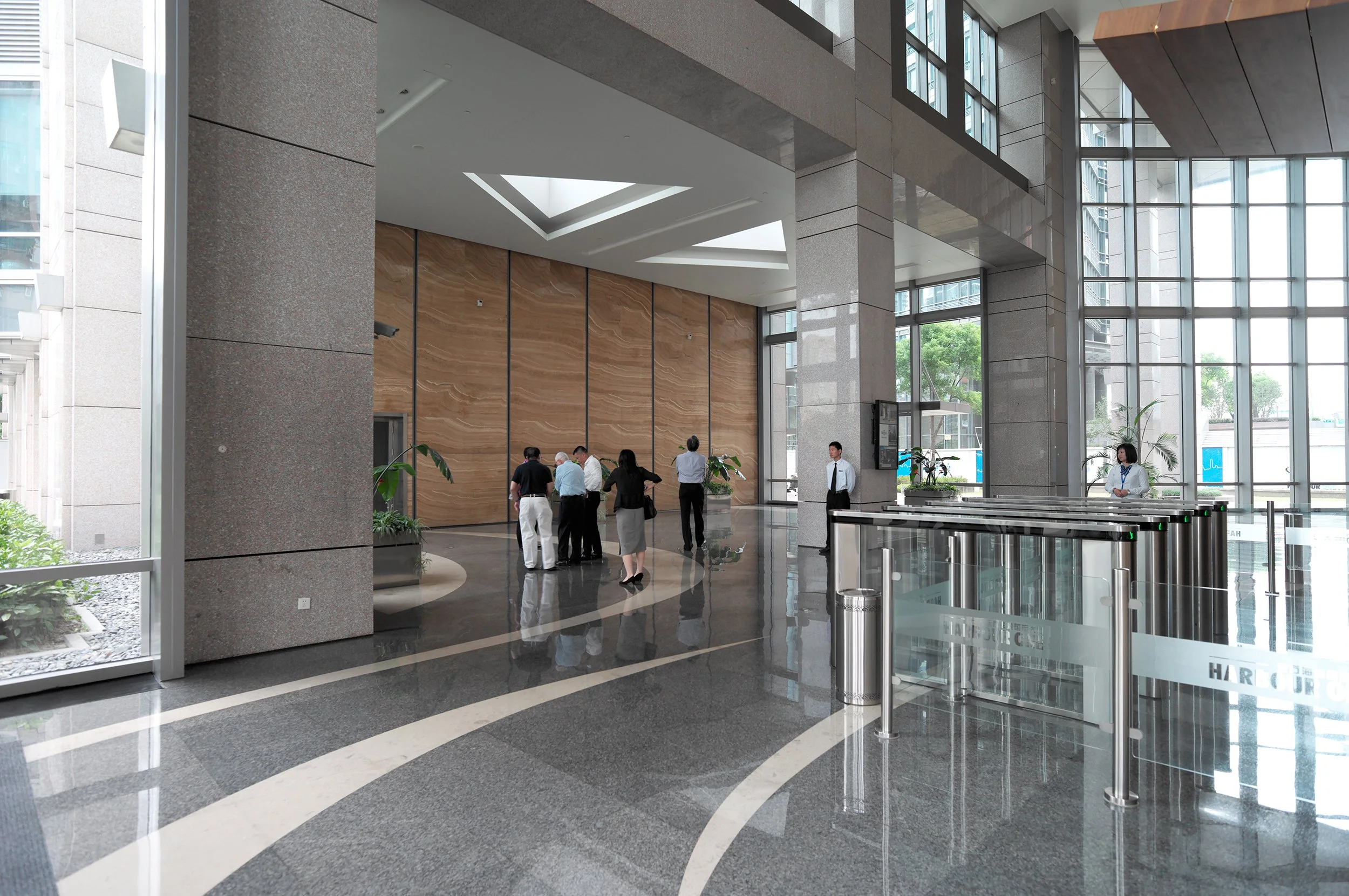 Modern office building lobby with large glass windows, group of people near wooden wall, and security turnstiles at entrance