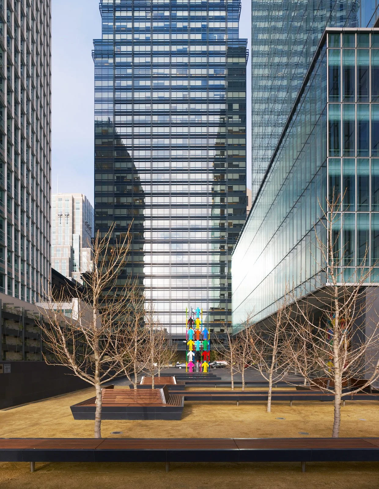 An outdoor urban space with leafless trees, benches, and a colorful sculpture of human figures, surrounded by tall modern glass buildings.