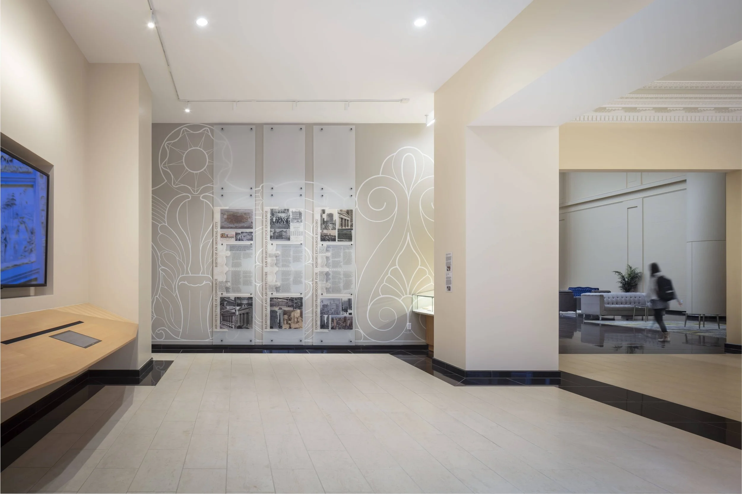 Interior of a modern building lobby with display panels, a TV, and seating area, featuring light-colored walls and flooring.