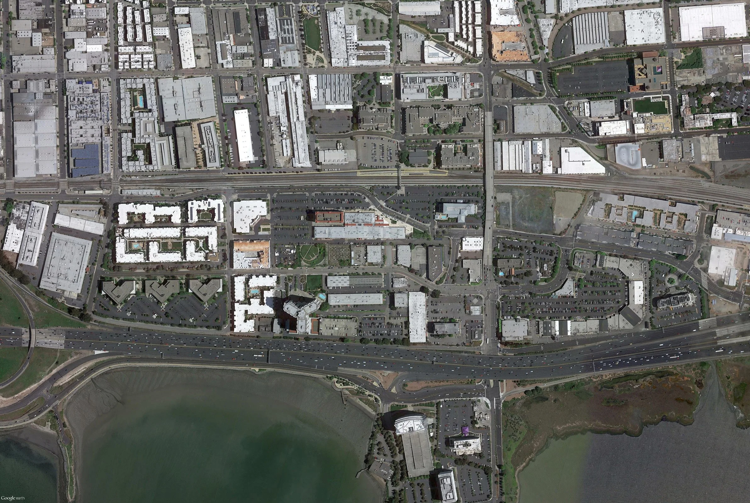 An aerial view of a city with buildings, roads, parking lots, a highway, and a body of water in the foreground.