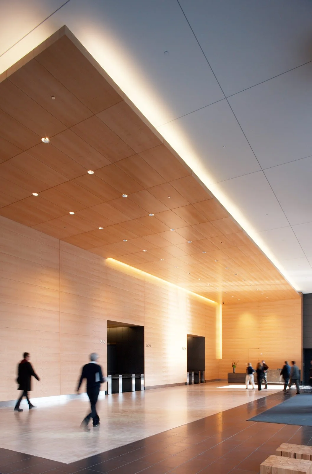 Interior view of a modern lobby with wooden walls and ceiling, featuring small ceiling lights and some people walking.