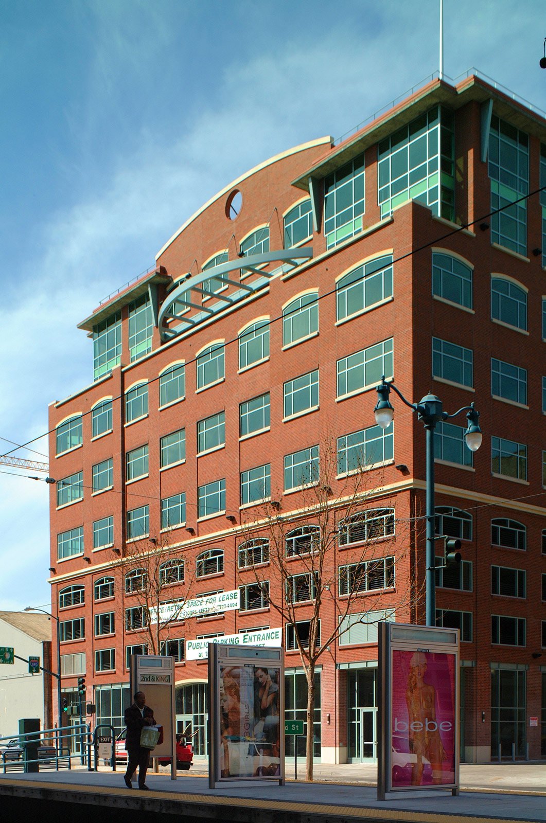 A multi-story brick building with large windows and modern architectural features, located at a bus stop with advertisements and a person waiting.