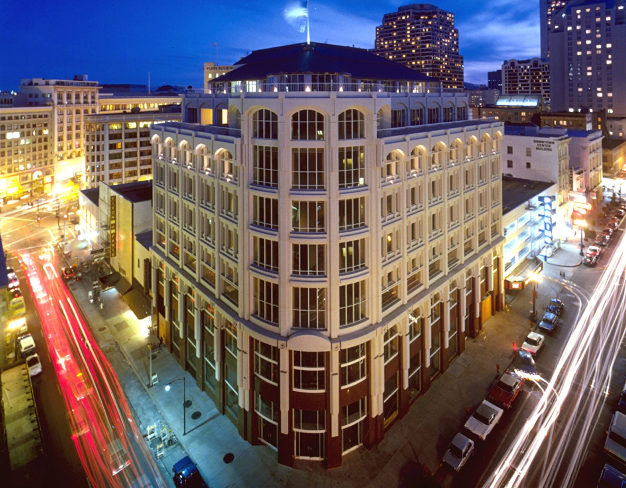 Nighttime cityscape featuring a multi-story building with large windows, traffic streaks from moving vehicles on the streets, and illuminated surrounding buildings.