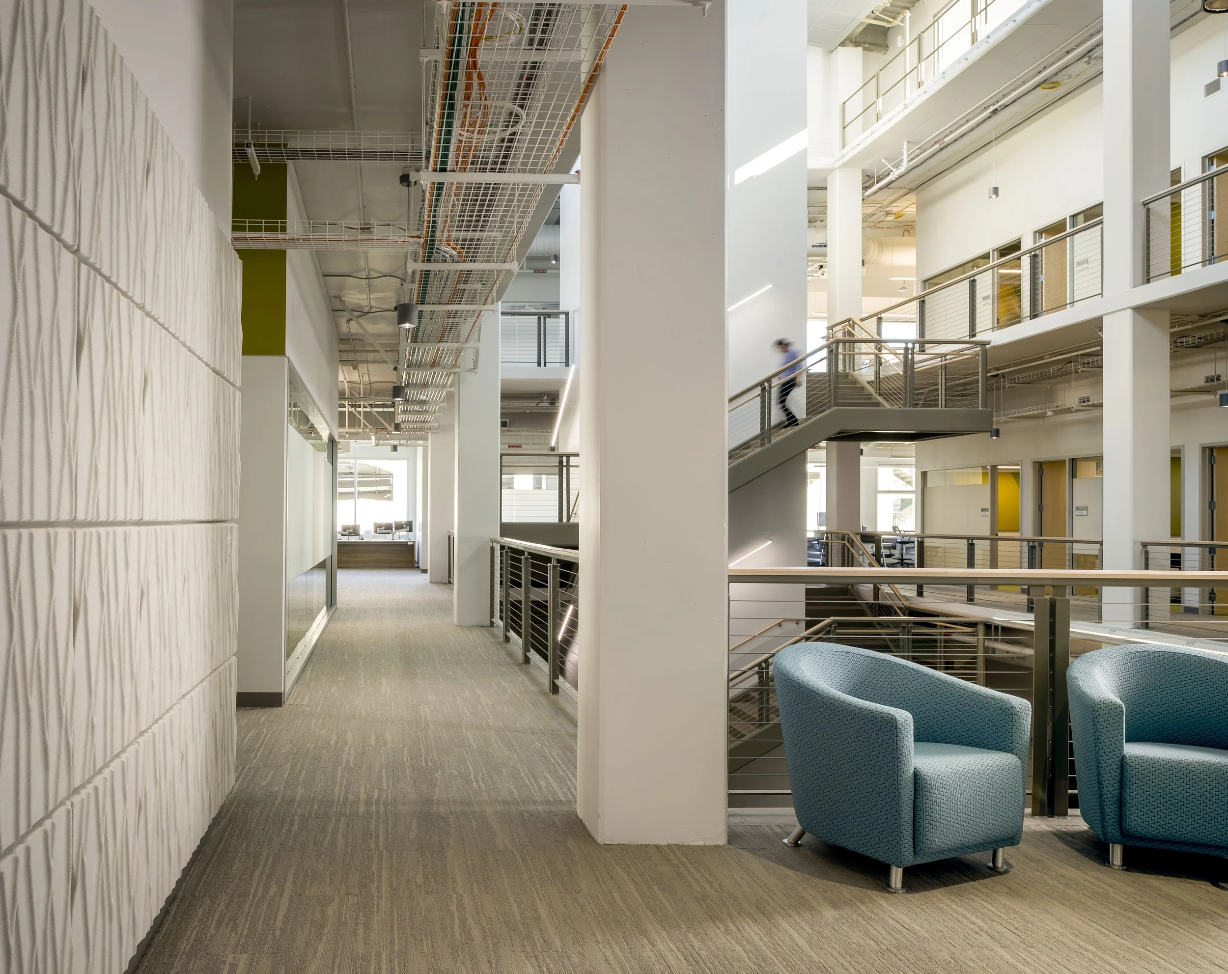 Modern office interior with open staircase, blue chairs, carpeted floor, and exposed ceiling pipes and wiring.