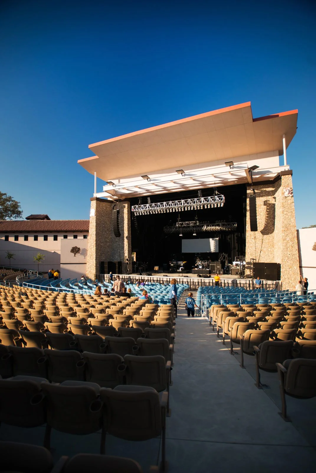 An outdoor concert stage with a large roof and lighting equipment set up, surrounded by rows of empty chairs, under a clear blue sky.