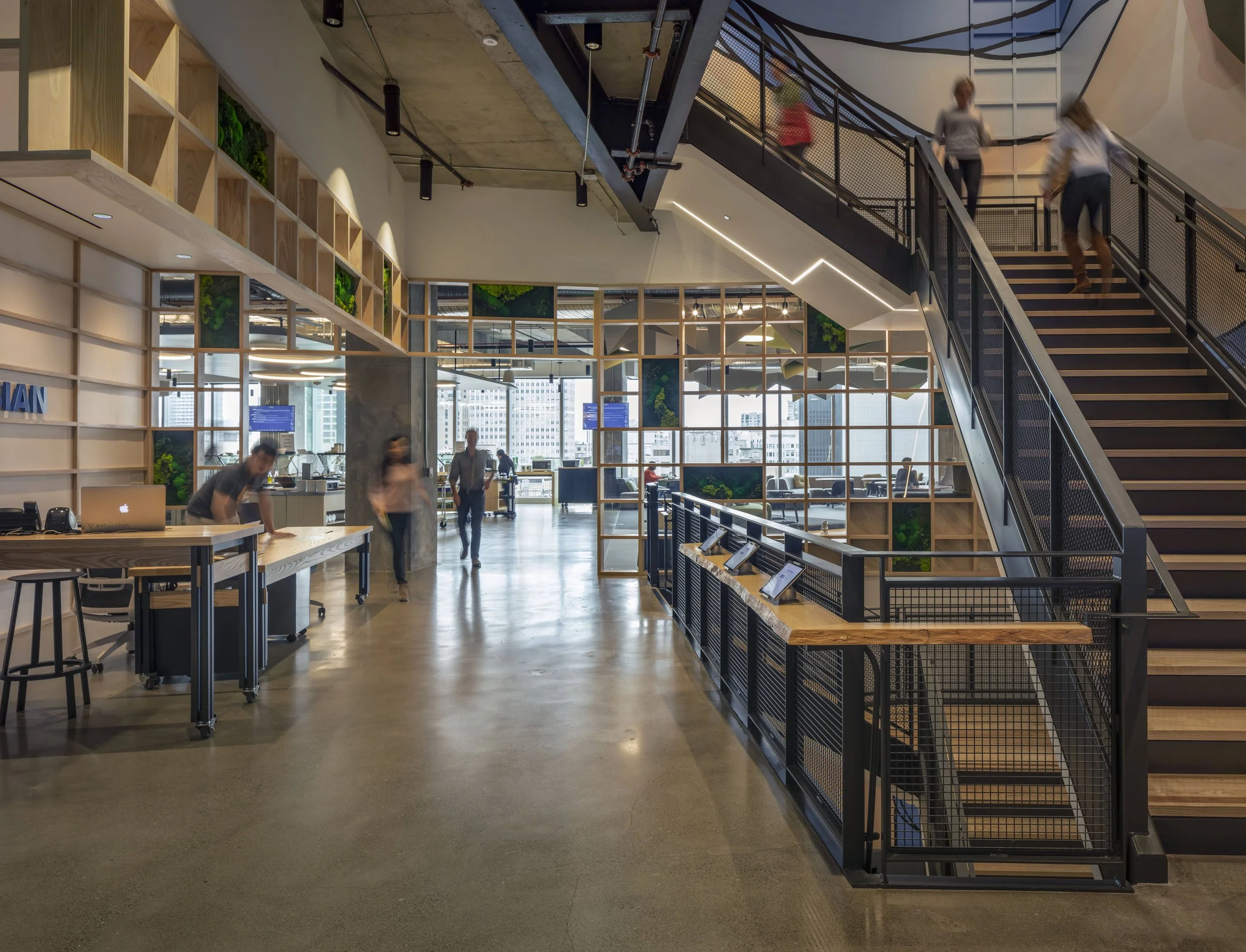 Modern office lobby with a staircase, glass walls, and people walking through.