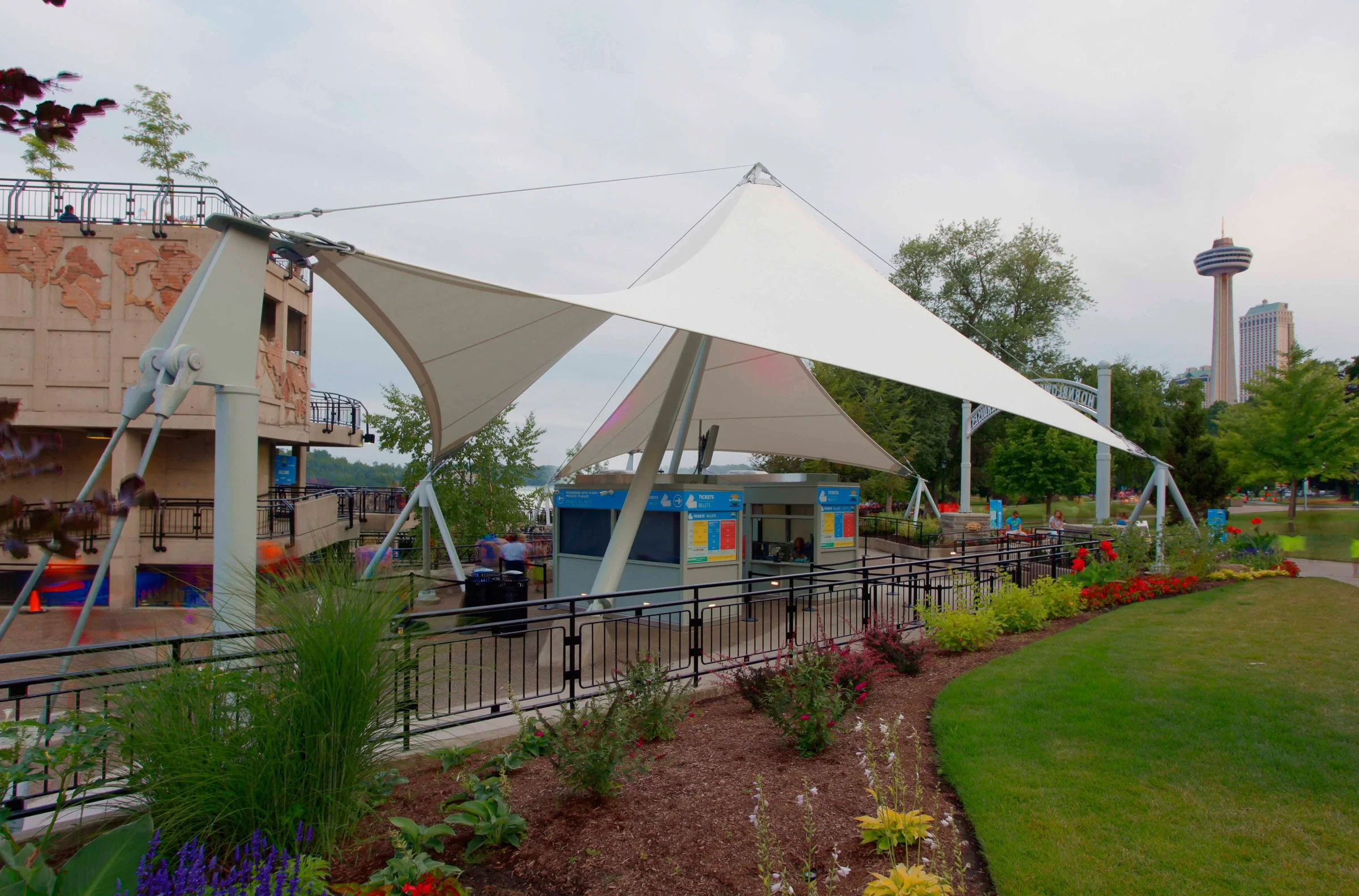 An outdoor amusement park entrance with a large white tensile fabric canopy, ticket booths, and a colorful flower garden in the foreground. A tower is visible in the background to the right.
