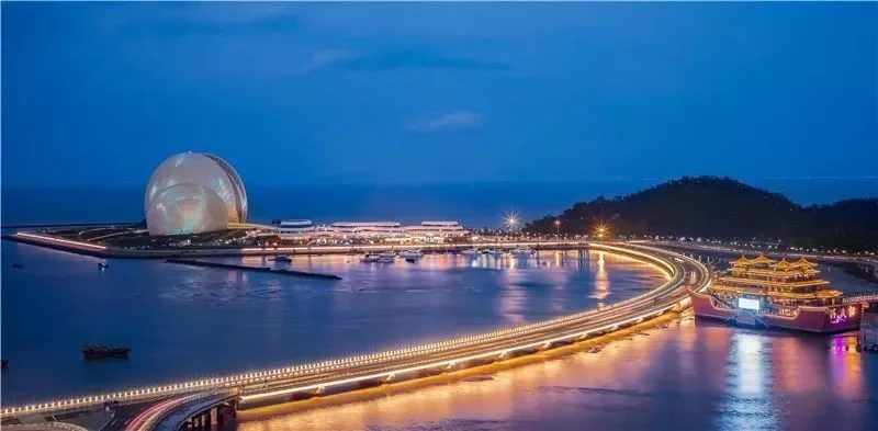 Night view of a cityscape with a large spherical building, a curved bridge with bright lights, and traditional Asian-style architecture near the water.