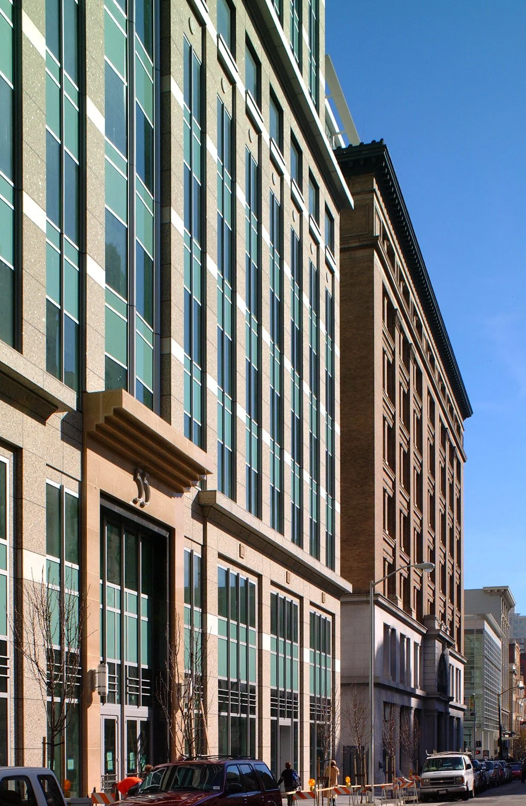 Photo of a city street with modern glass and stone buildings, some trees, parked cars, and a few pedestrians, under a clear blue sky.