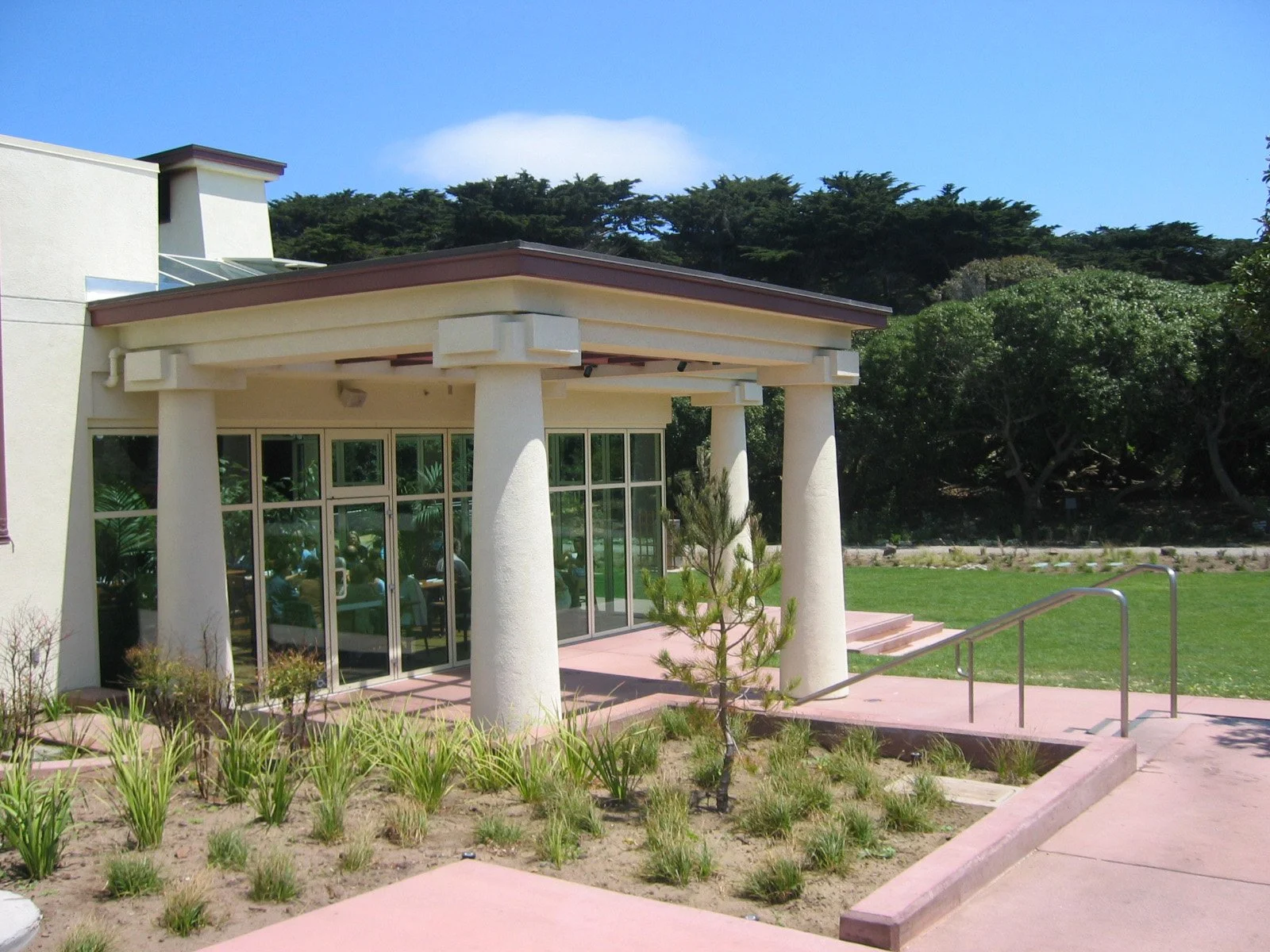 Exterior view of a building with large glass doors and windows, surrounded by a landscaped garden with small plants and a young tree. There is a ramp for accessibility and lush green trees in the background under a blue sky.