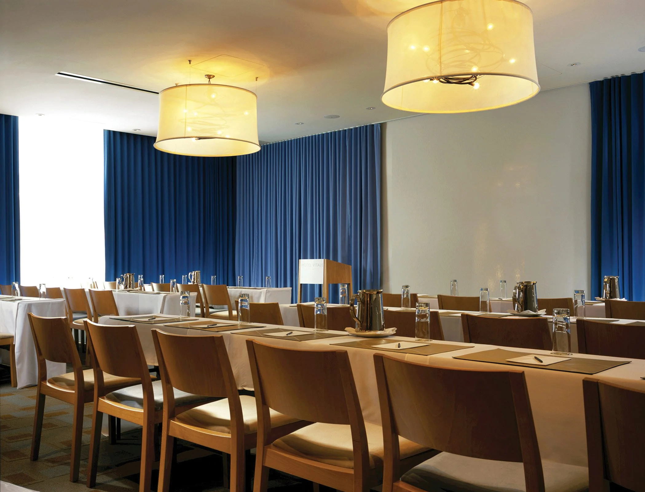 Conference room with chairs, tables, notepads, water glasses, pitchers, and large ceiling lights. Blue curtains cover the windows.