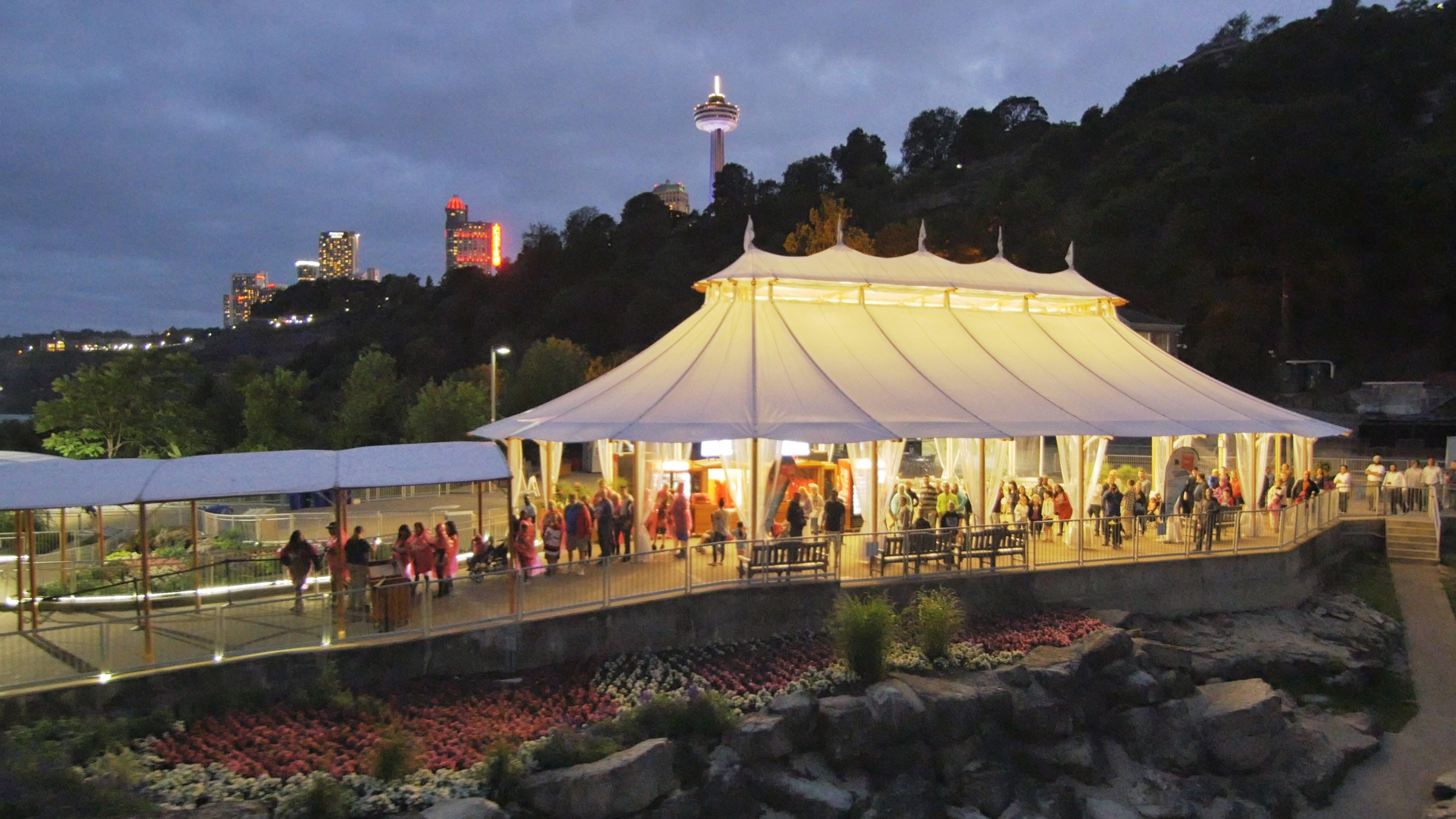 A lit tent with a pointed roof and many people inside and outside, surrounded by a walkway, with city lights and a tower in the background, at dusk.