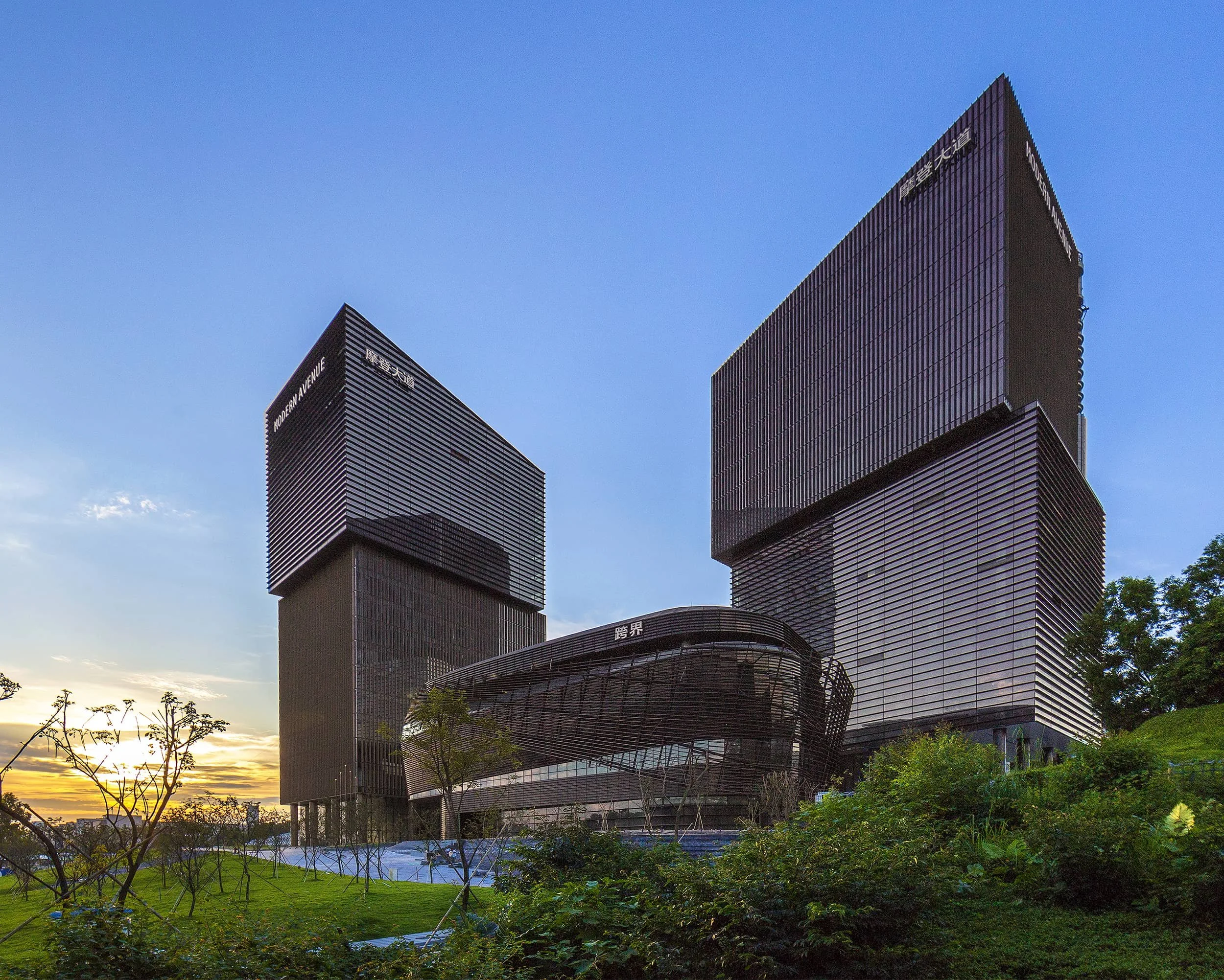 Two modern black skyscrapers with horizontal lines and the signs 'MODERN AVENUE' and 'FUKUOKA' at sunset, with green foliage and trees in the foreground.
